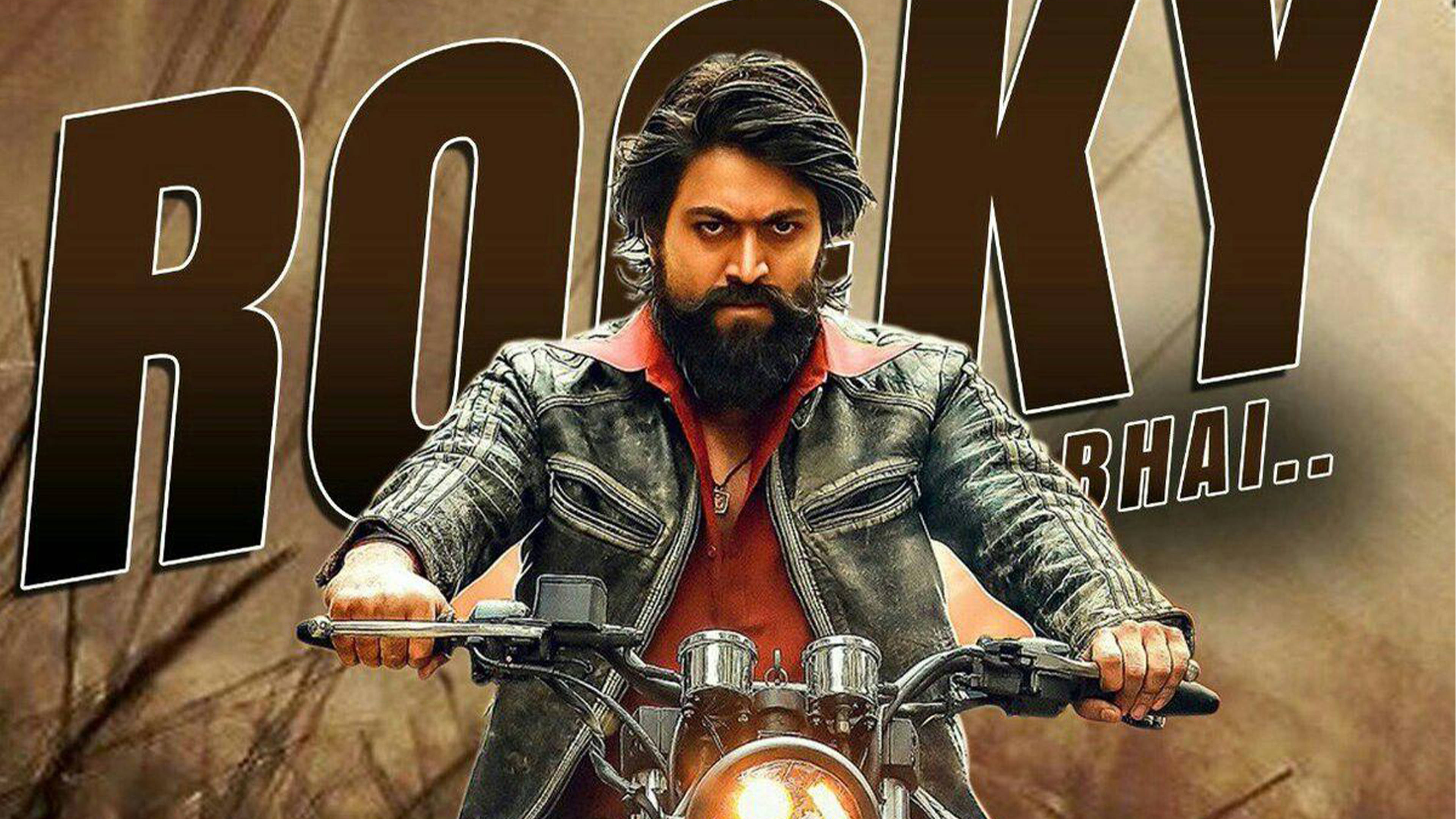 kgf 4K 桌布 壁紙 — 一個留鬍鬚的男人穿著皮夾克騎摩托車，身後有 'ROCKY BHAI..' 文字。