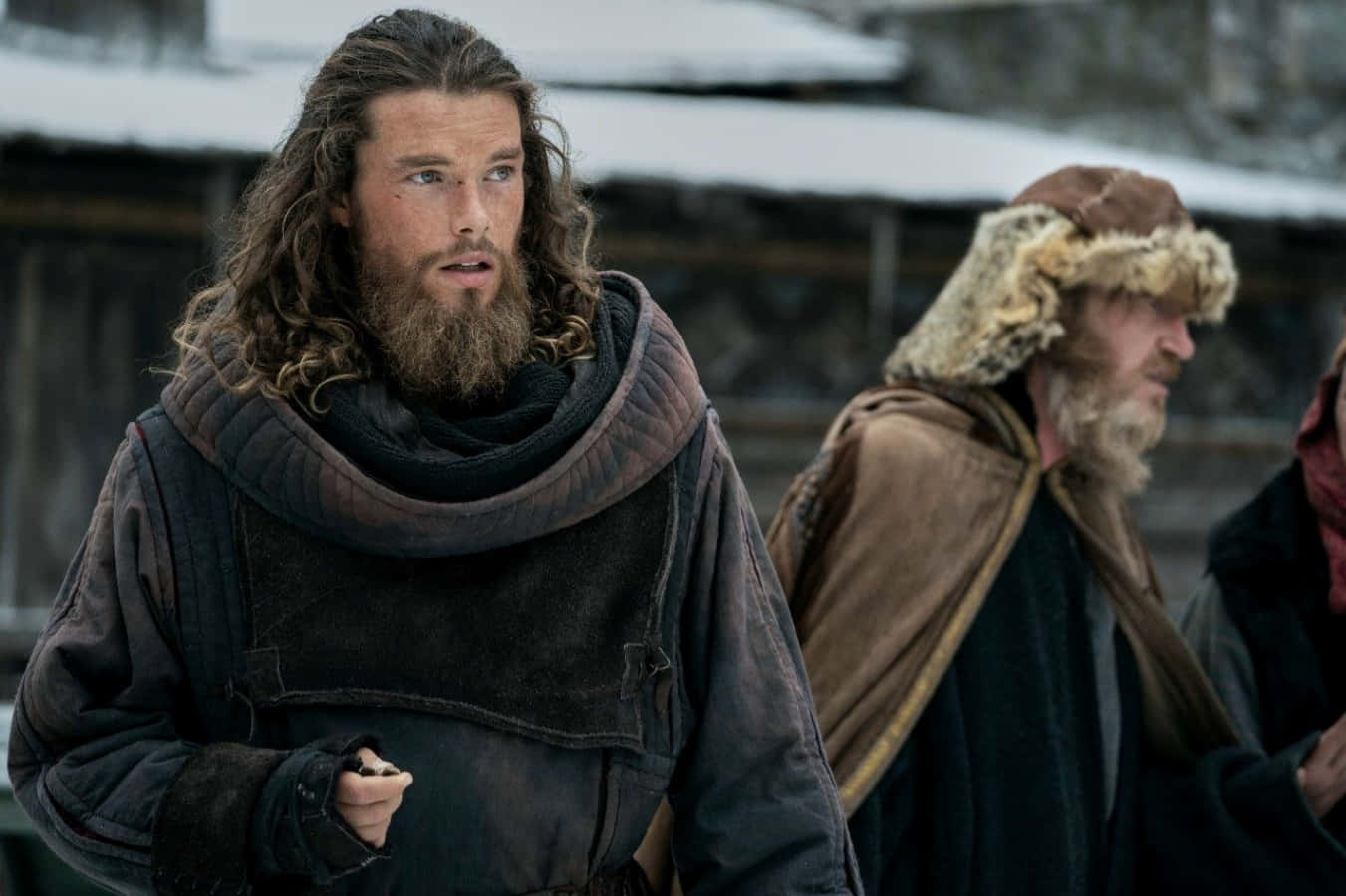 Vikings 桌布 壁紙 — 一個留鬍鬚的男人穿著粗獷的冬天衣服，在一個下雪的村莊警覺地站著，旁邊還有另一個人。