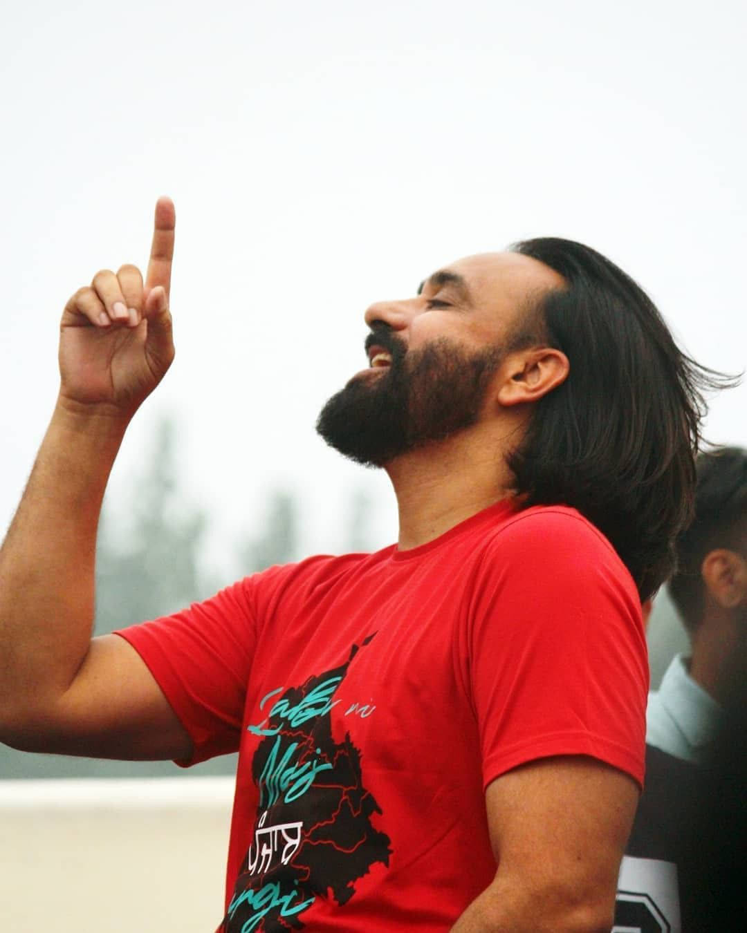 Indian 桌布 壁紙 — 一個留鬍鬚的男人穿著紅色T恤，在模糊的戶外背景下，帶著燦爛的笑容愉快地向上指著。