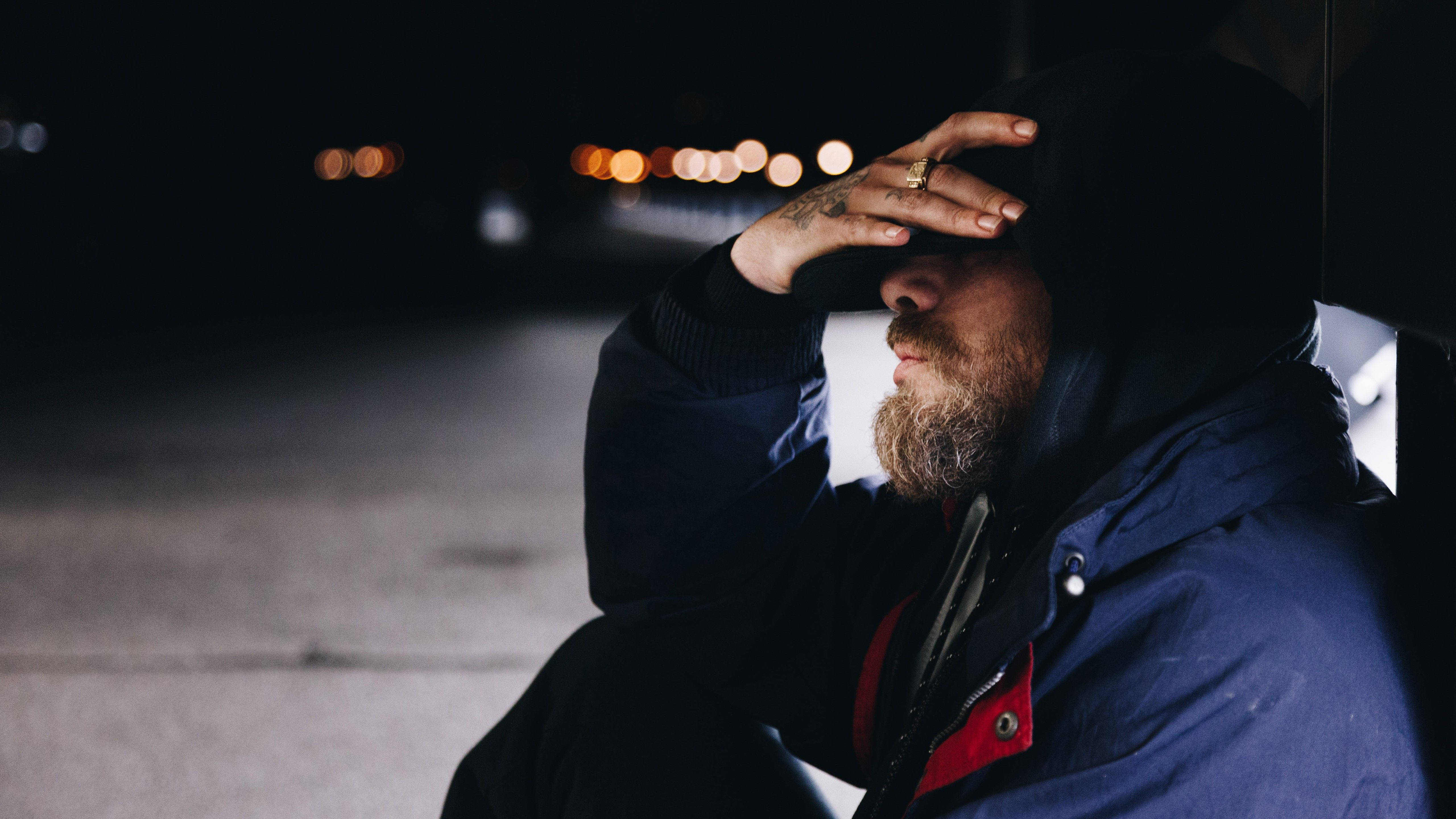 Alone Boy 桌布 壁紙 — 一個留鬍鬚的男人穿著連帽外套，夜晚獨自坐著，手放在額頭上，背景是模糊的路燈。