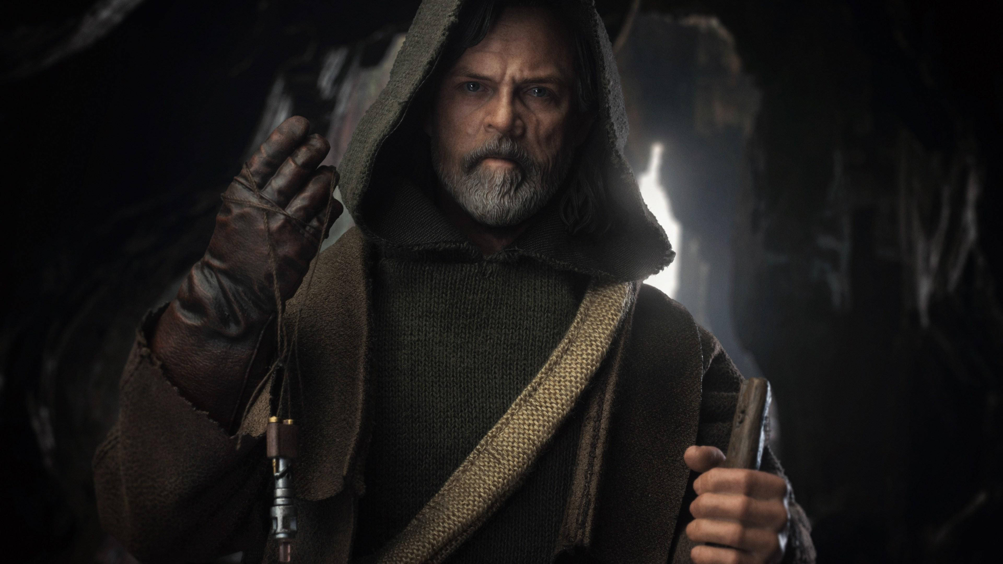 Star Wars Luke Skywalker 4K 桌布 壁紙 — 一個留鬍鬚的男人穿著連帽長袍，在昏暗的洞穴中握著光劍柄，身後有明亮的光芒。