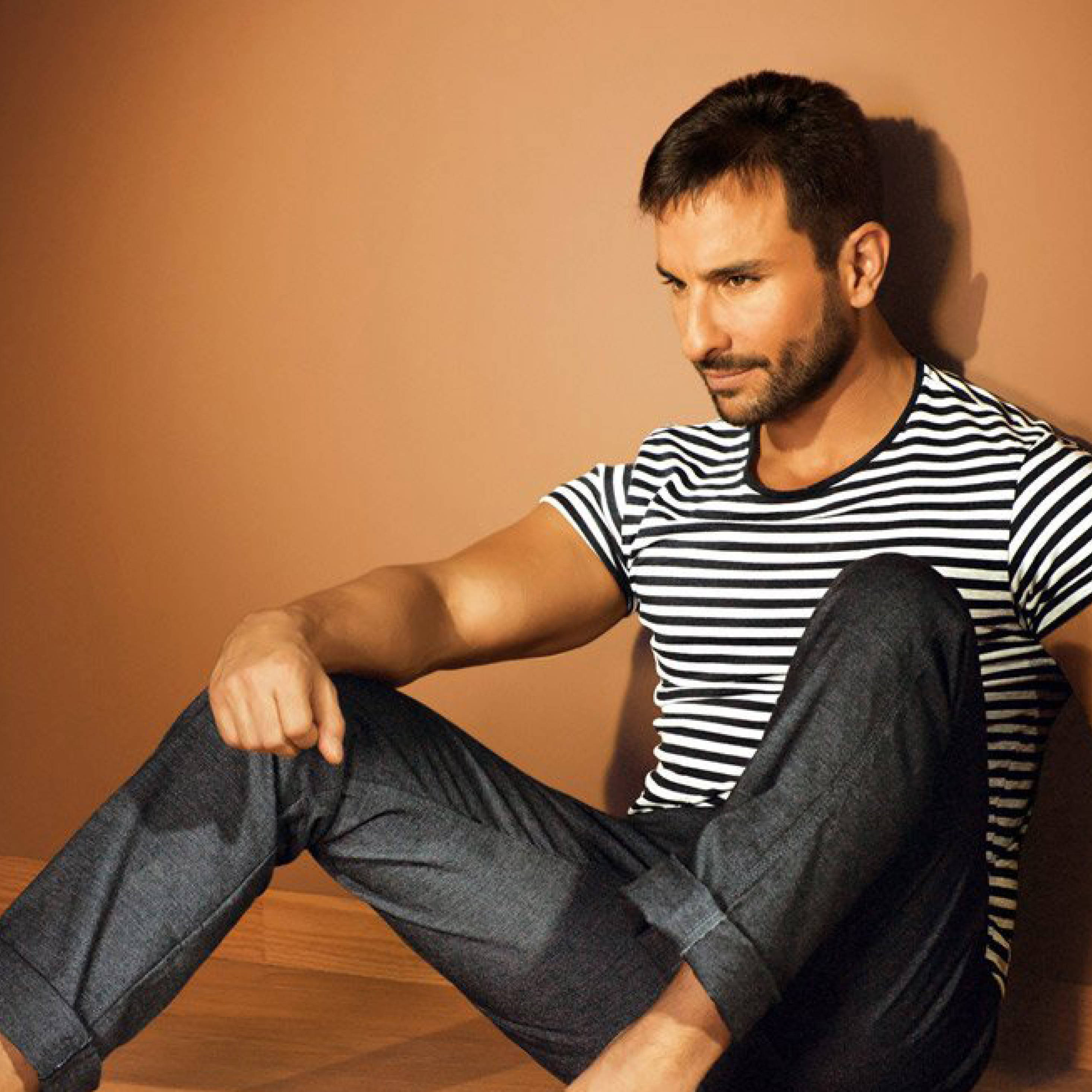 Saif Ali Khan 桌布 壁紙 — 一個留鬍鬚的男人穿著黑白條紋衫和捲起褲管的牛仔褲，隨意地靠著暖色調的牆壁坐著。