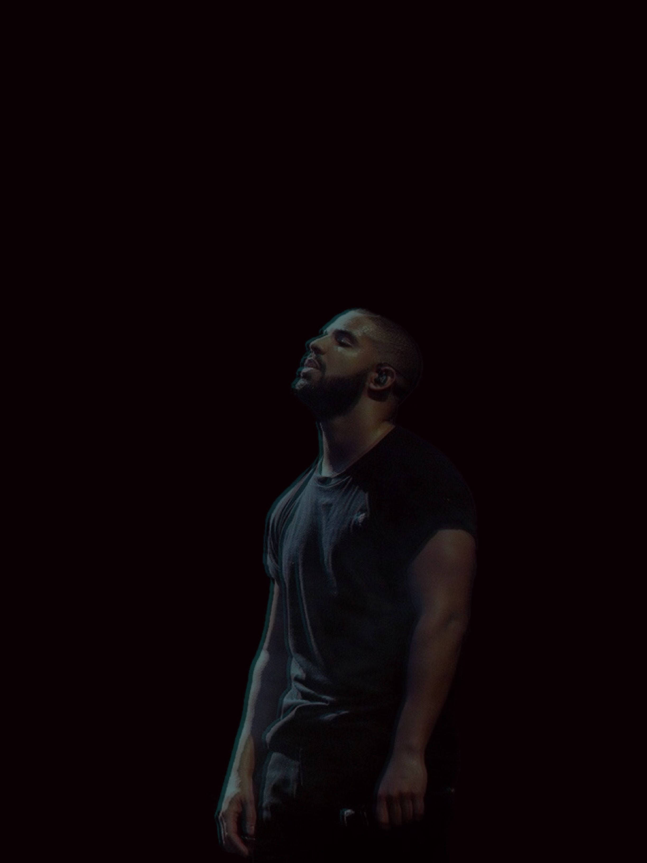 Drake 桌布 壁紙 — 一個留鬍鬚的男人站在近乎黑暗的環境中，他的臉被柔和的光線照亮，抬頭仰望，表情沉思。