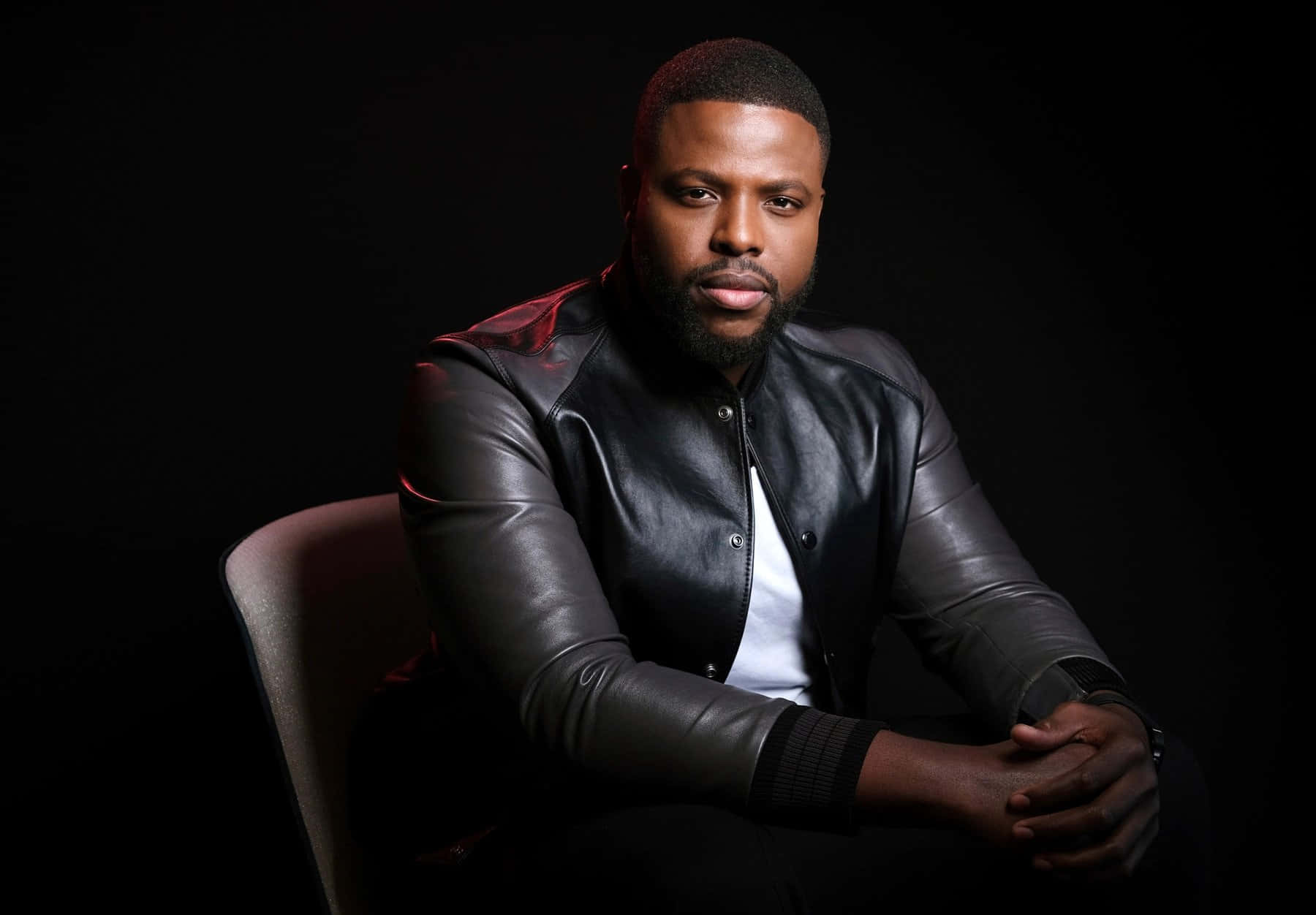 Winston Duke 桌布 壁紙 — 一個留鬍鬚的男人自信地坐在椅子上，穿著黑色皮夾克，背景黑暗帶有紅色燈光點綴。