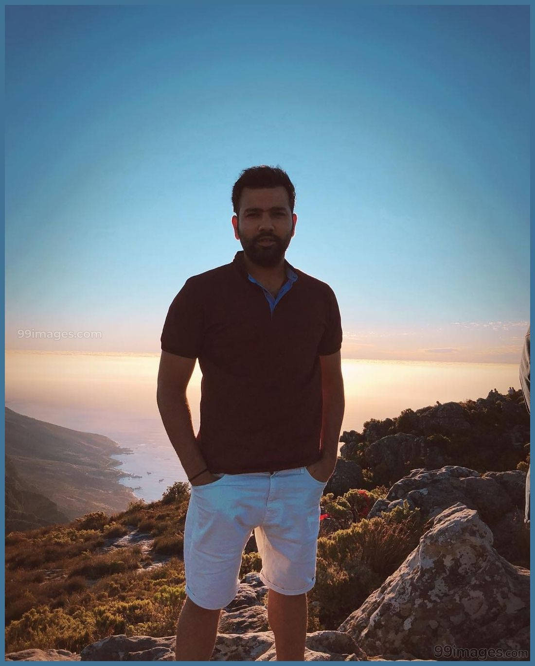 Rohit Sharma 桌布 壁紙 — 一個留鬍鬚的男人自信地站在岩石峭壁上，俯瞰著溫暖金色光線的海岸日落。