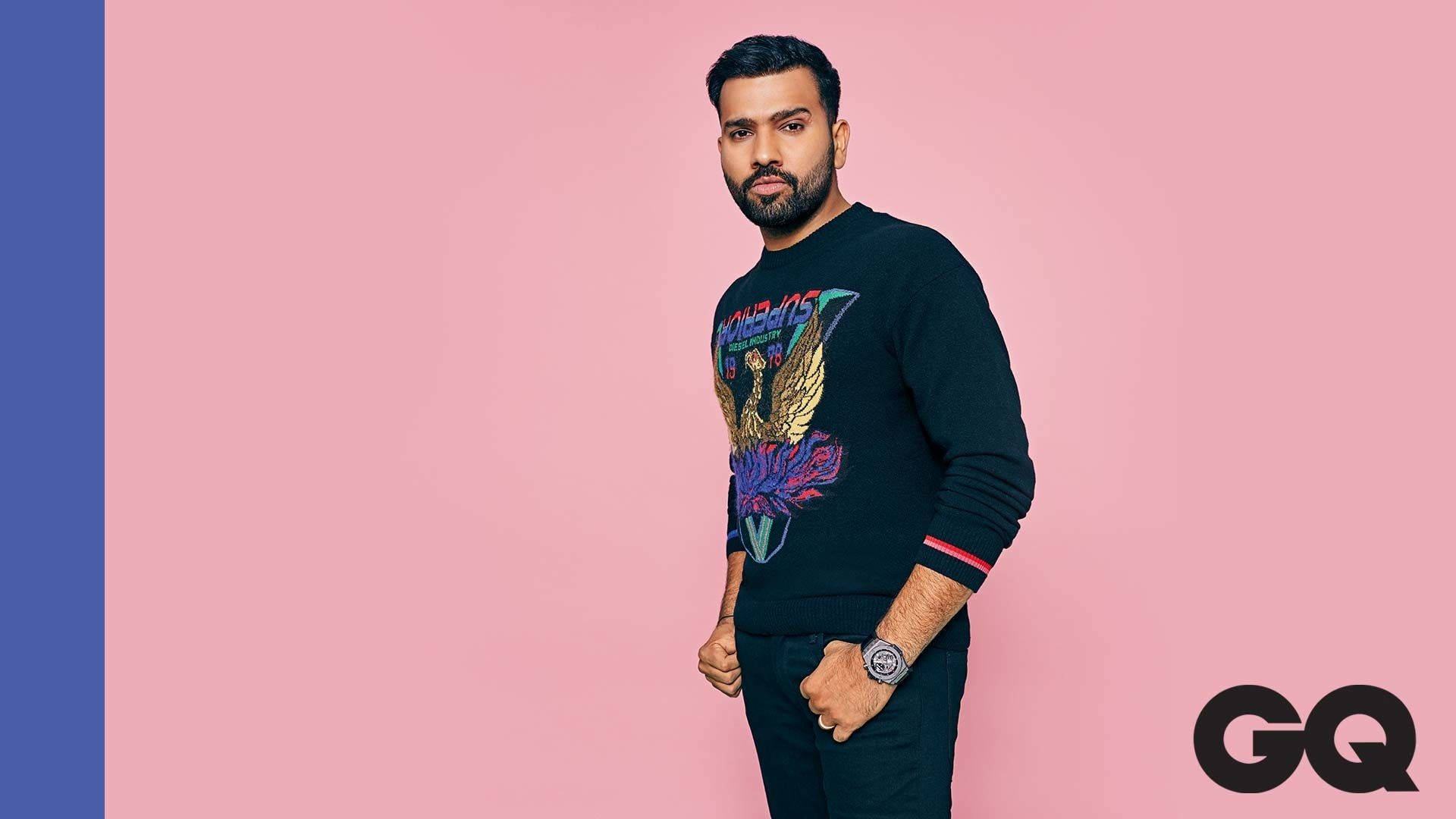 Rohit Sharma 桌布 壁紙 — 一個留鬍鬚的男人自信地站在粉紅色和藍色背景前，身穿黑色毛衣，上面有色彩繽紛的鷹圖案。