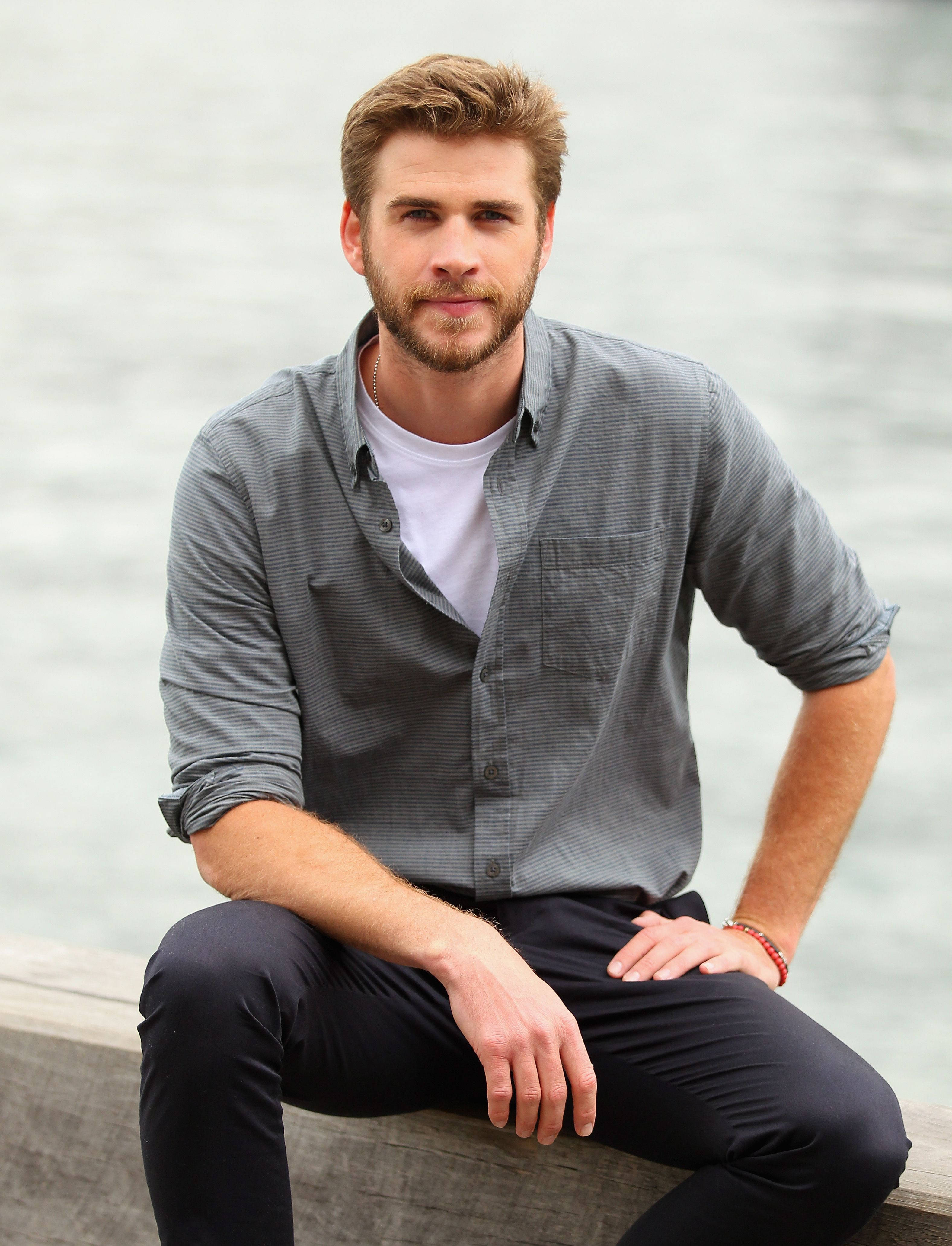 liam 桌布 壁紙 — 一個留鬍鬚的男人隨意地坐在平靜水邊的混凝土邊緣，穿著灰色鈕扣襯衫和深色褲子。