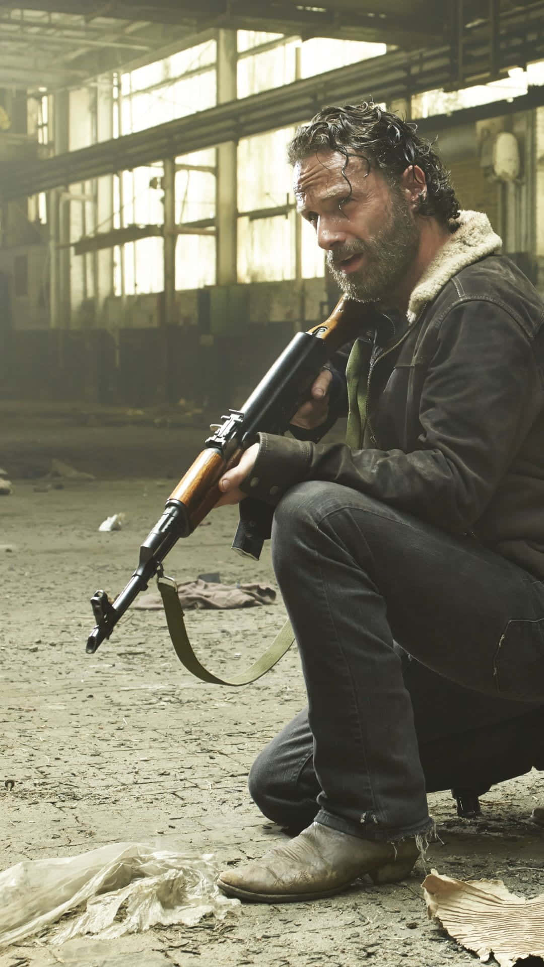 The Walking Dead iPhone 桌布 壁紙 — 一個留鬍鬚的粗獷男人在破舊的倉庫中蹲下，手持突擊步槍，表情陰沉。