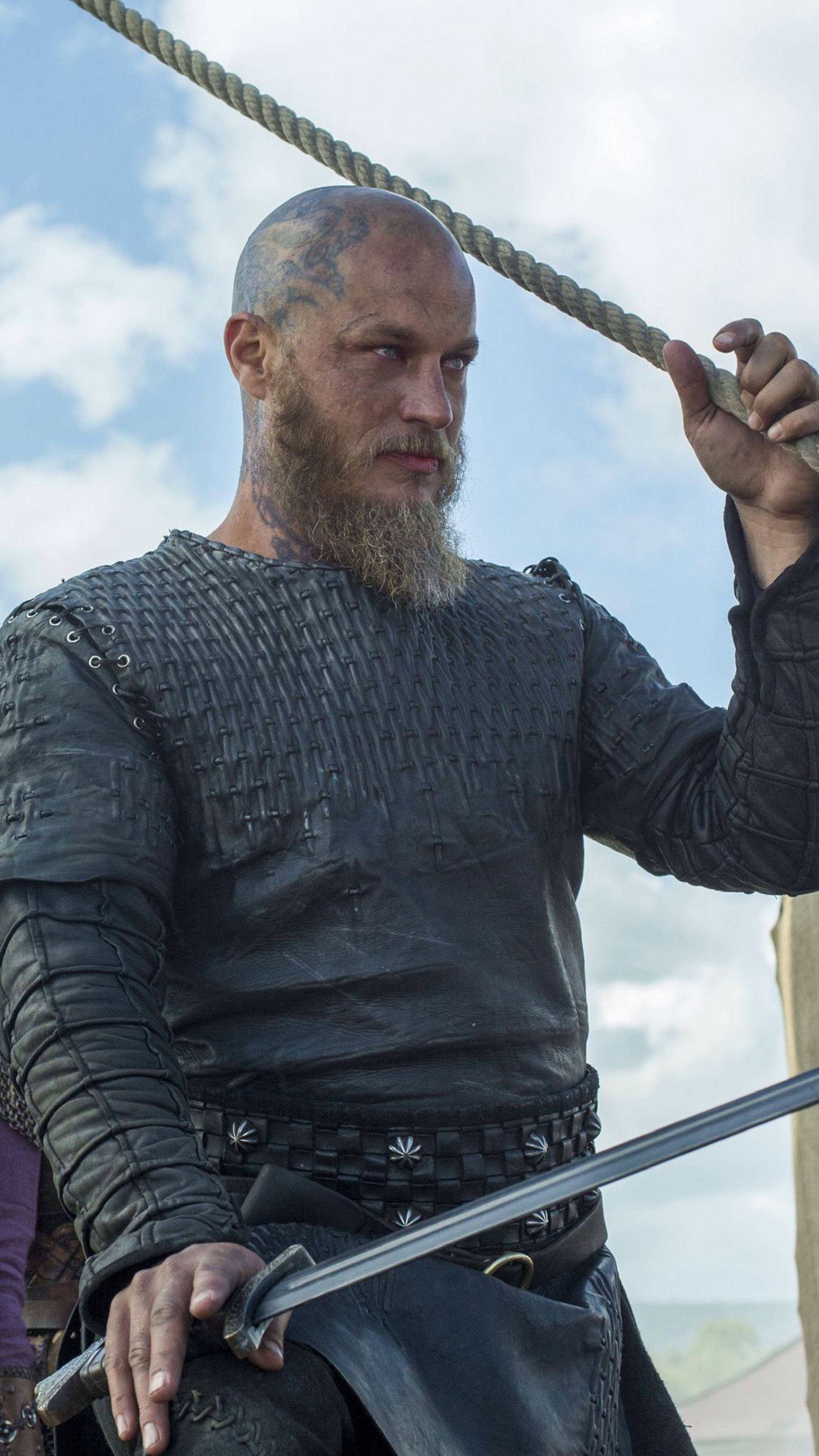 Vikings 桌布 壁紙 — 一個留鬍鬚的維京戰士身穿深色皮甲，在陰雲密佈的天空下抓著繩索和劍。