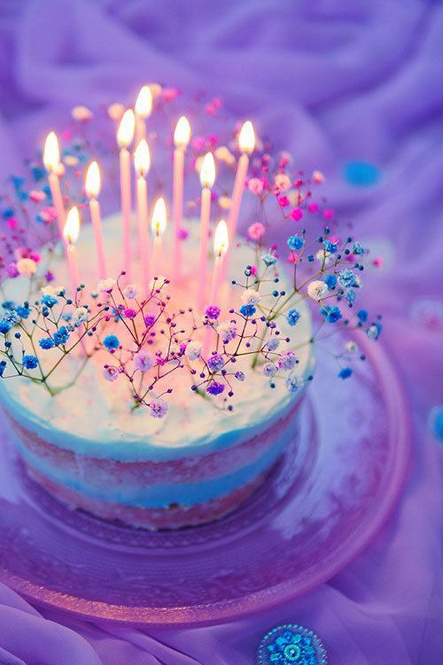 Birthday Cake 桌布 壁紙 — 一個異想天開的生日蛋糕，帶有發光的蠟燭和柔和的滿天星花朵，放在紫色布料背景上。