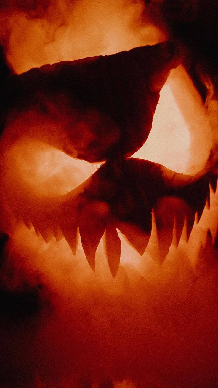 Scary Halloween iPhone 桌布 壁紙 — 一個發光、帶有鋒利牙齒和熾熱橙色光芒、在黑暗煙霧中顯得威嚇的傑克南瓜燈面孔。