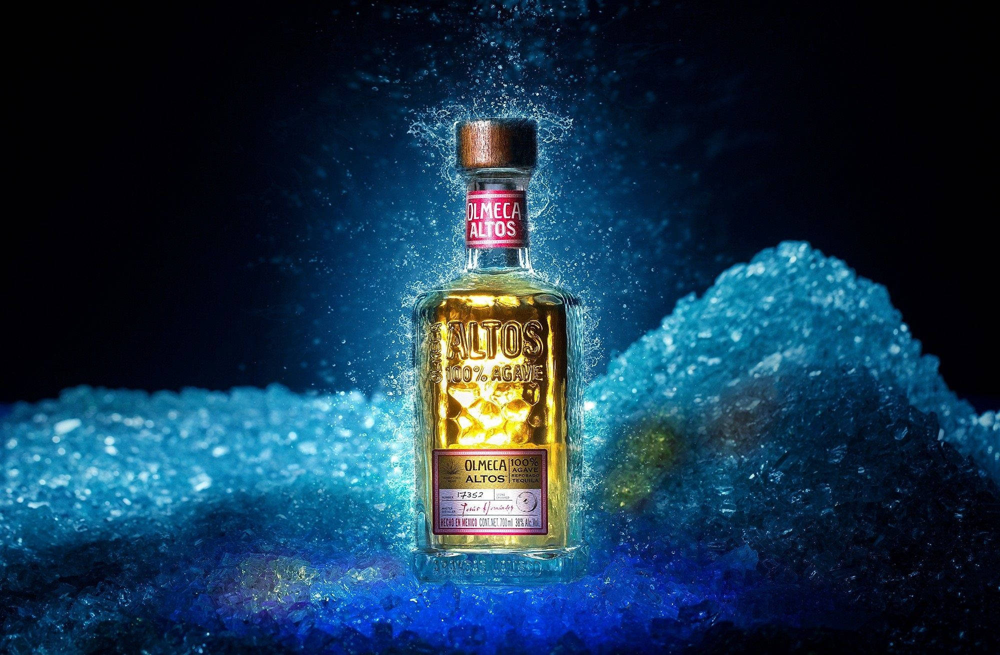alcohol 桌布 壁紙 — 一個發光的 Olmeca Altos 龍舌蘭酒瓶，在深色背景下，立於冰藍色液體飛濺之中。