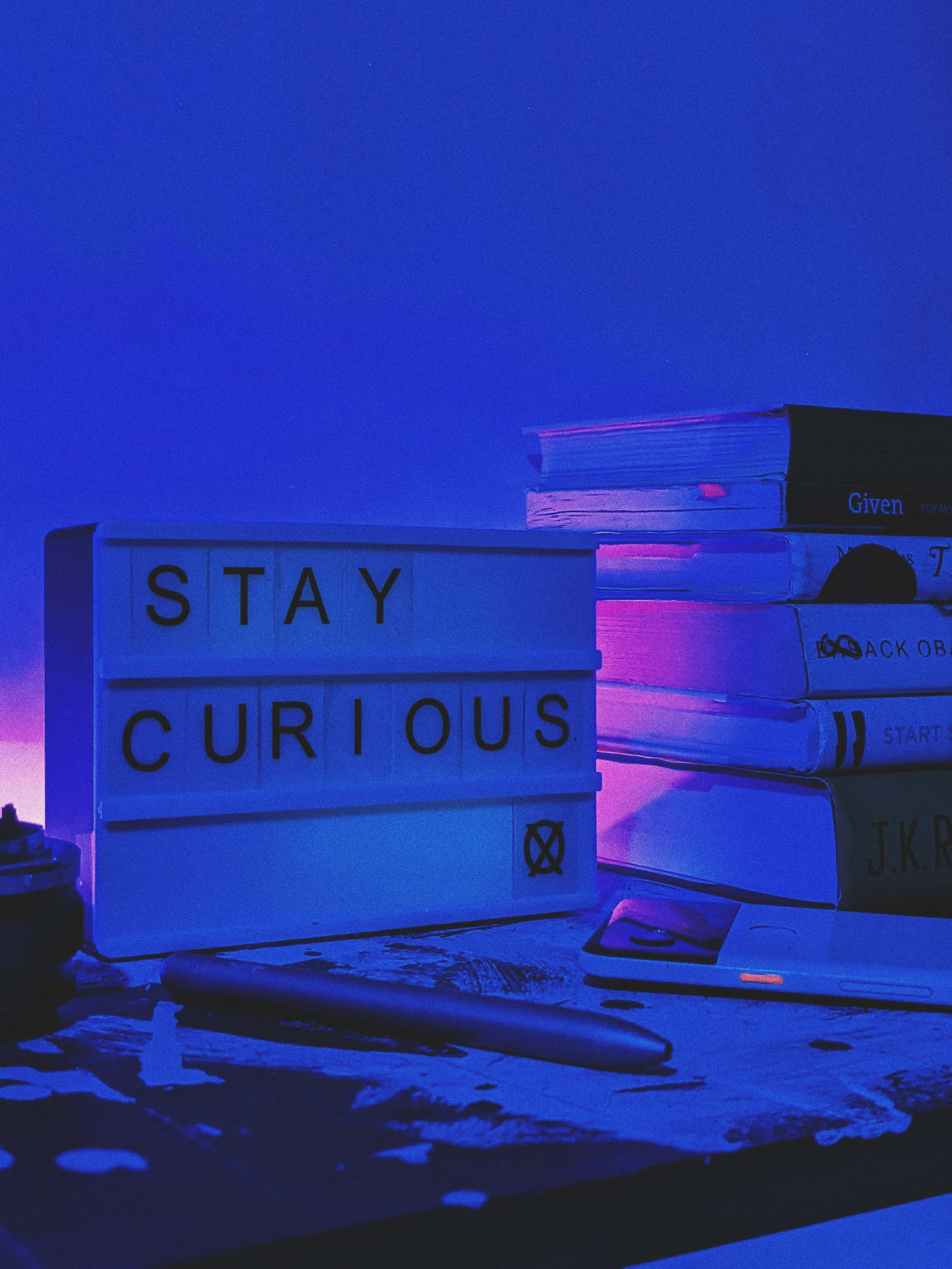 手機 桌布 壁紙 — 一個發光的「STAY CURIOUS」標誌沐浴在藍色和紫色燈光中，旁邊放著一疊書和一支筆，置於有紋理的桌子上。