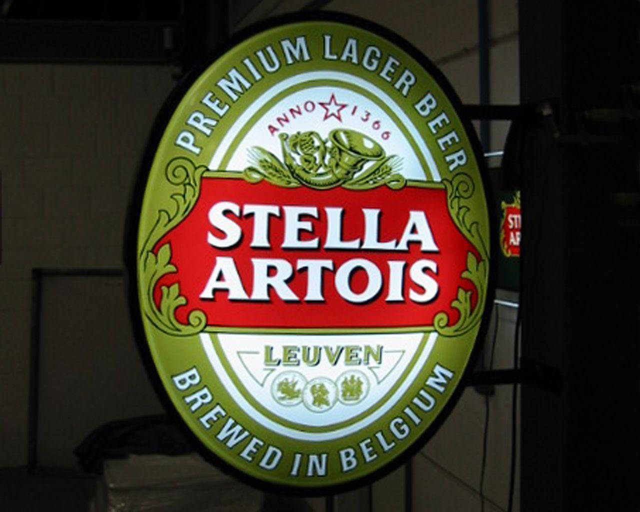 飲料 桌布 壁紙 — 一個發光的 Stella Artois 啤酒招牌，帶有綠色、紅色和白色，掛在燈光昏暗的酒吧裡。