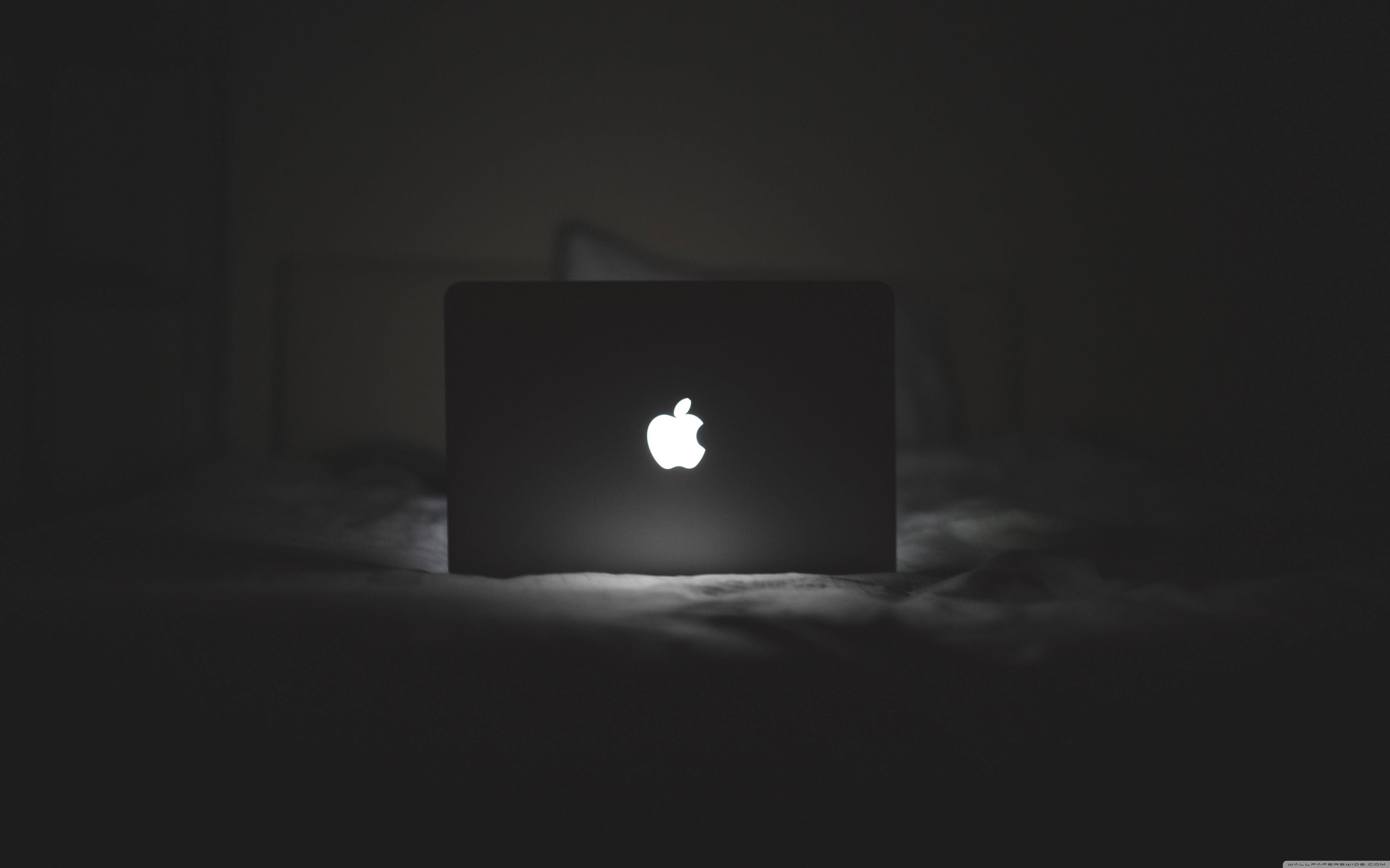 MacBook 動態圖片 桌布 壁紙 — 一個發光的Apple標誌在關閉的筆記型電腦上，獨自放在昏暗房間的深色床上。