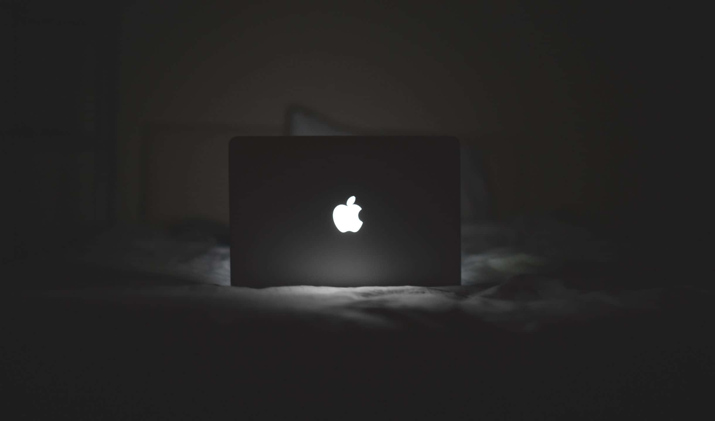 蘋果 桌布 壁紙 — 一個發光的Apple標誌在關閉的筆記型電腦上，獨自放在昏暗房間的深色床上。
