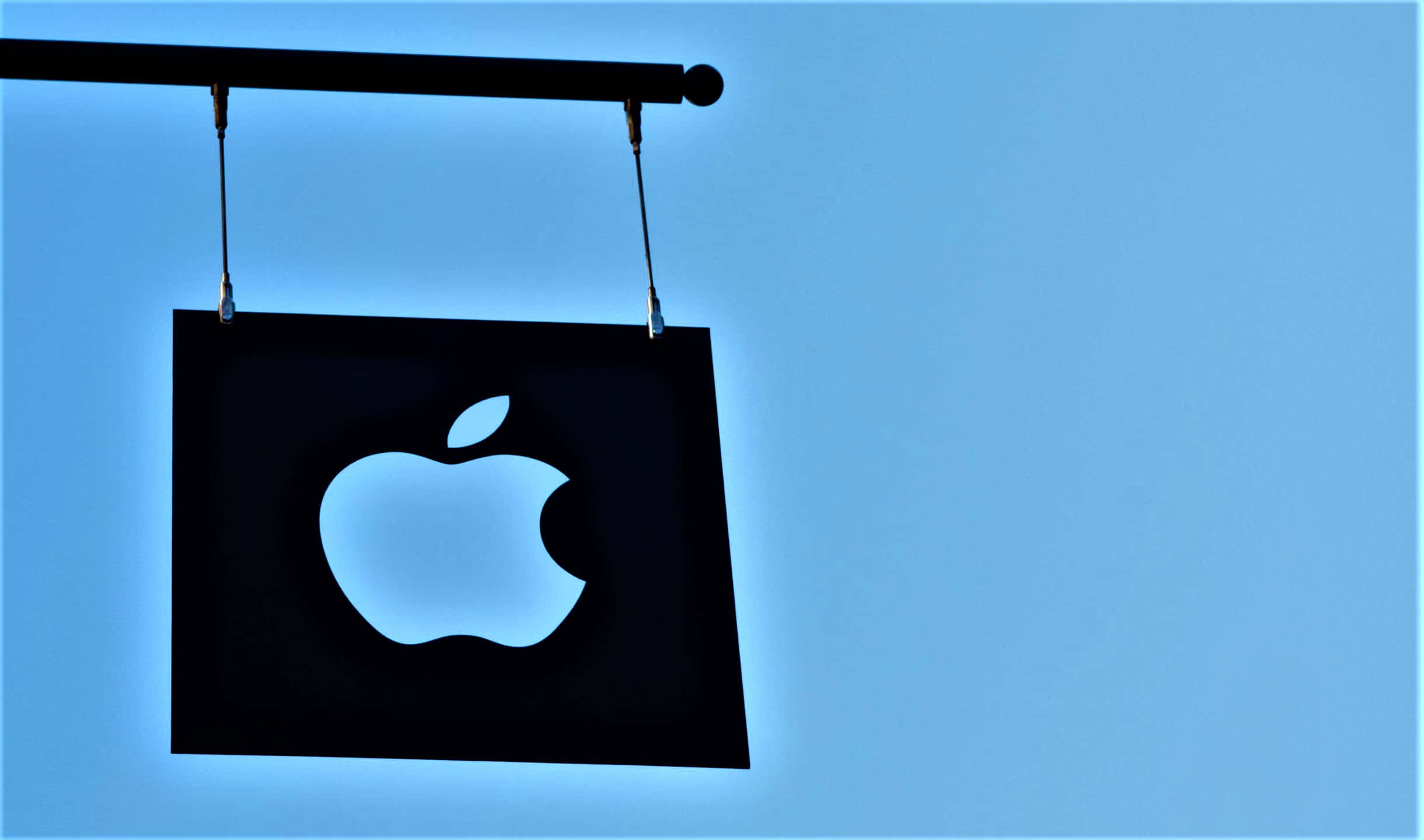 Blue Sky 桌布 壁紙 — 一個發光的Apple標誌招牌懸掛在清澈的藍天上，從內部散發出柔和的光芒。