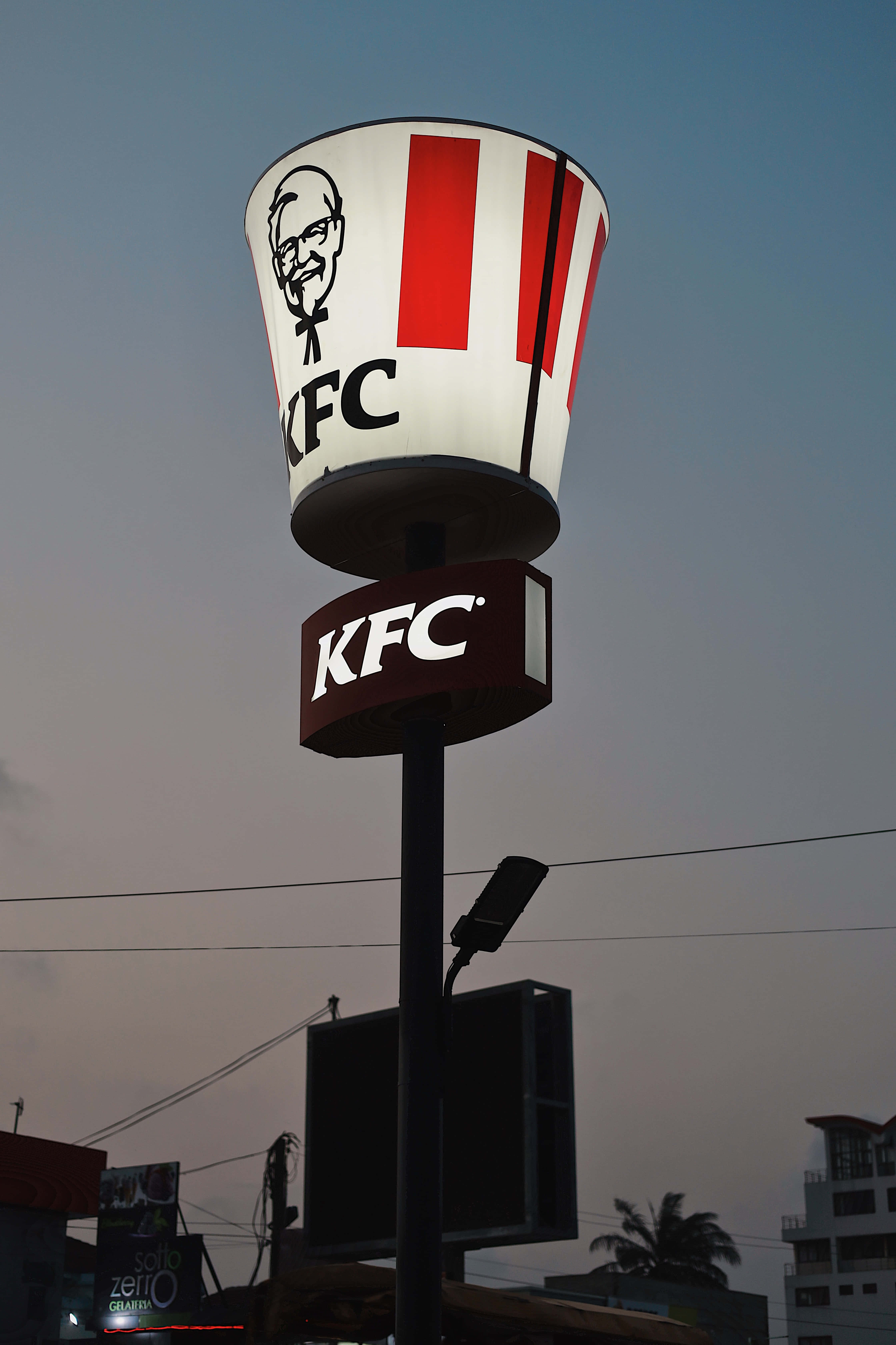 Kfc 桌布 壁紙 — 一個發光的KFC桶形招牌聳立在黃昏的天空下，背景有城市建築物和棕櫚樹。