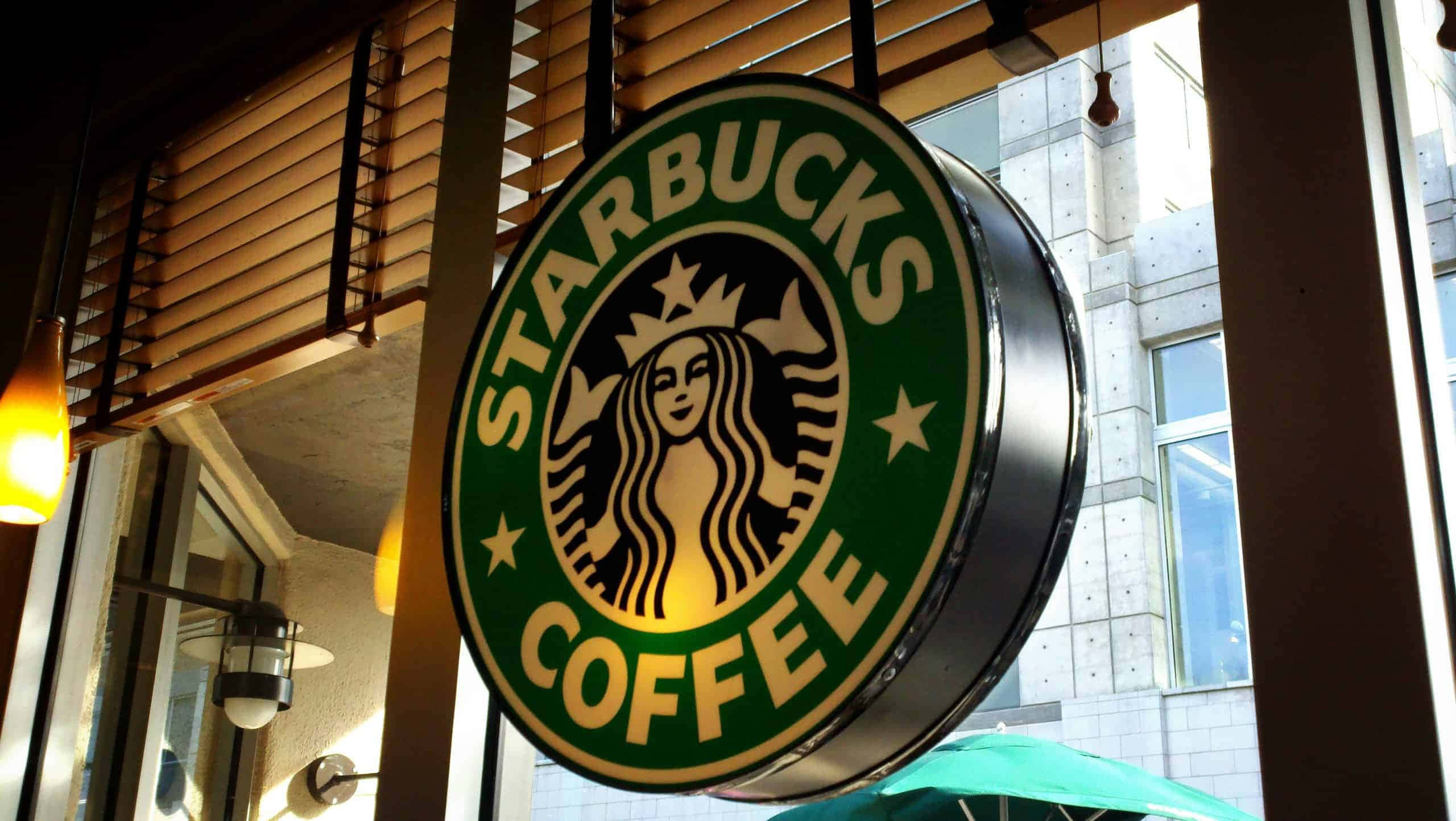 星巴克 桌布 壁紙 — 一個發光的Starbucks咖啡標誌掛在店鋪外，店內有溫暖的燈光和木製百葉窗。