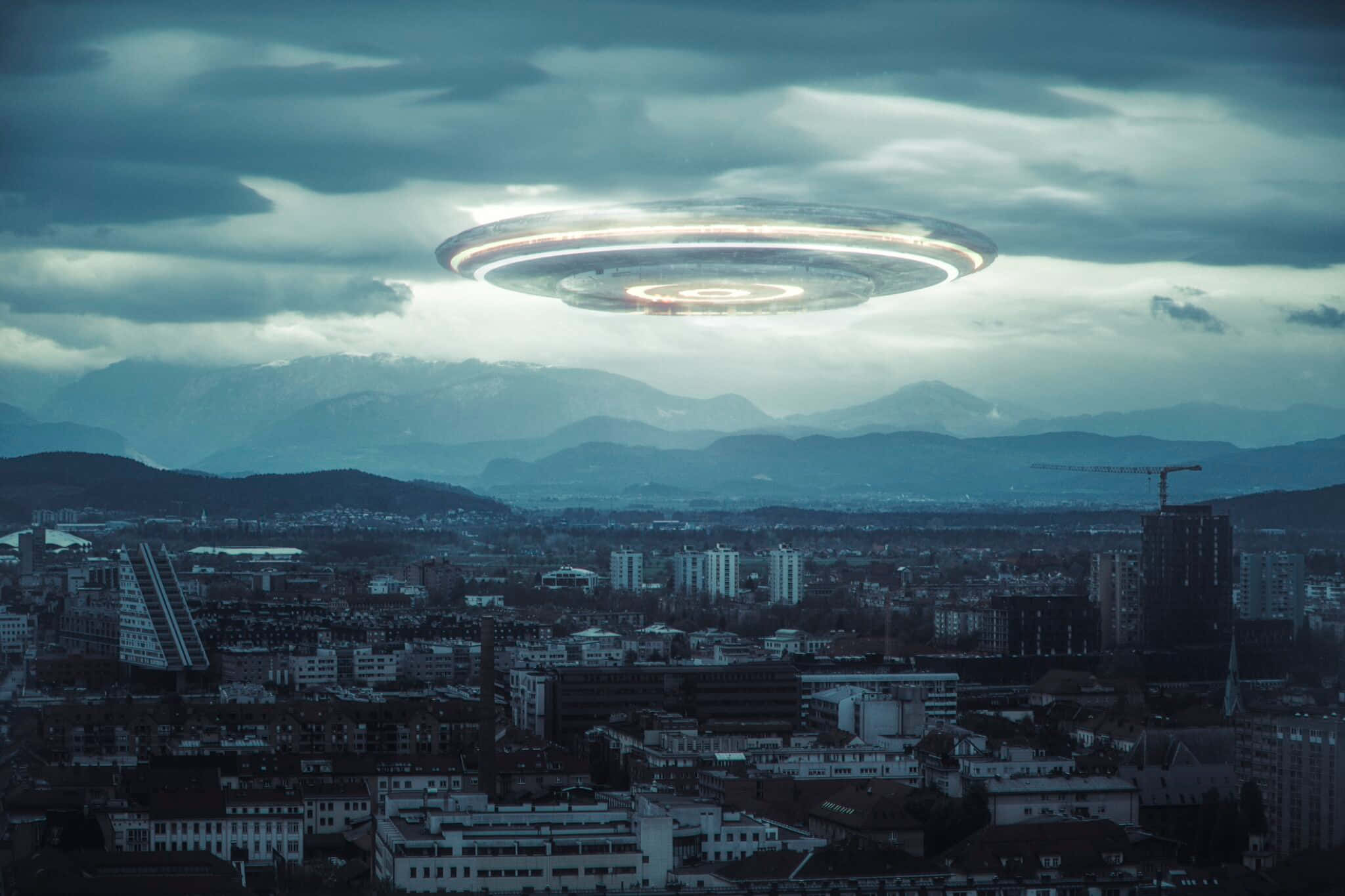 Real Ufo 桌布 壁紙 — 一個發光的UFO懸浮在廣闊的城市景觀上空，背景有山脈，天空多雲。