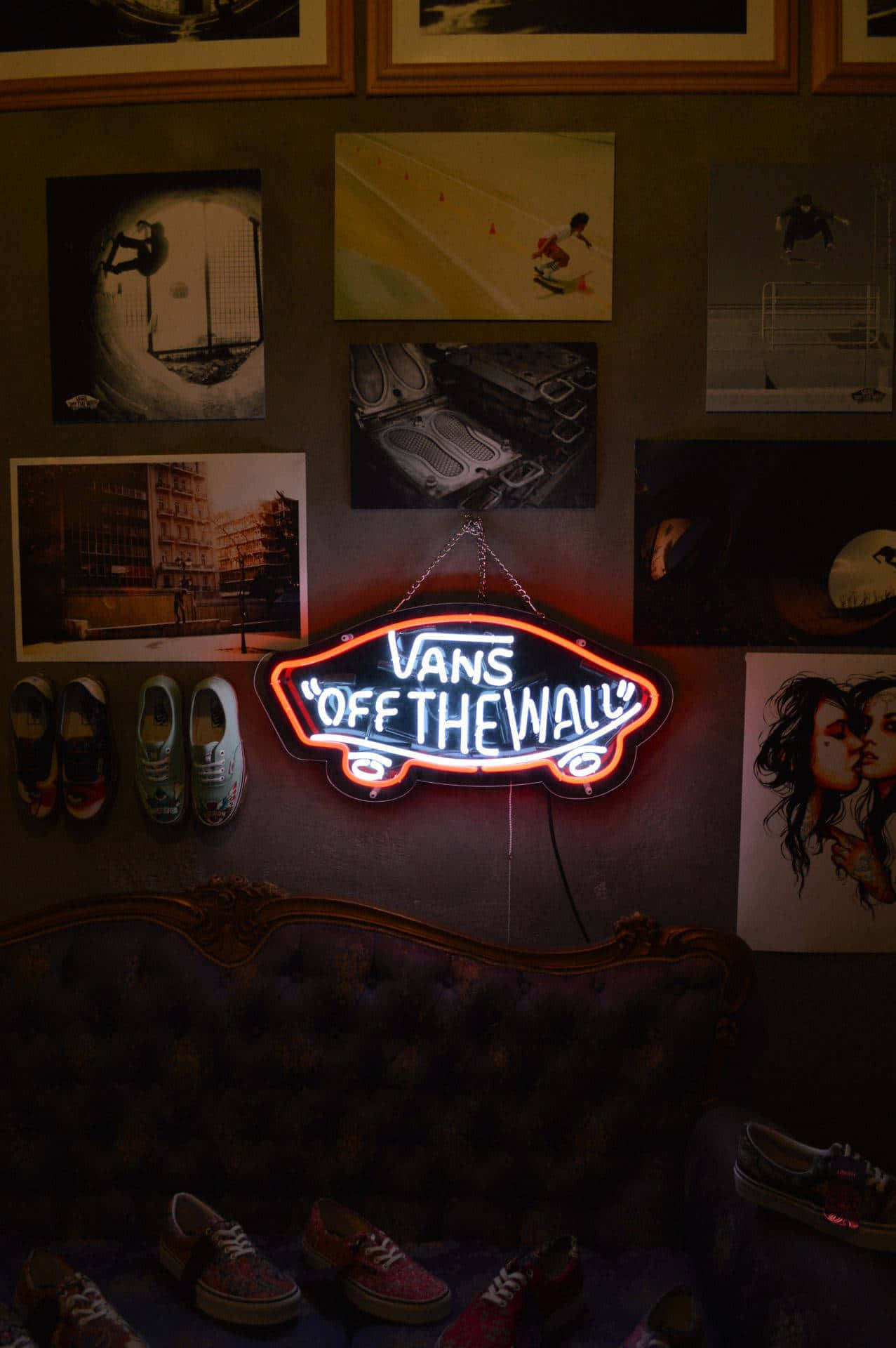 Vans 桌布 壁紙 — 一個發光的Vans Off The Wall霓虹招牌掛在天鵝絨沙發上方，房間光線昏暗，裝飾著滑板海報和色彩繽紛的運動鞋。