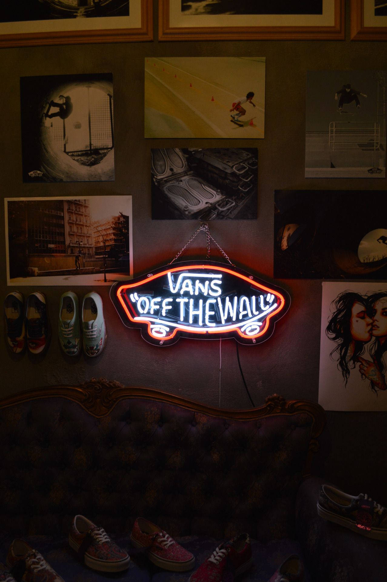 Vans 桌布 壁紙 — 一個發光的Vans霓虹燈牌，形狀像滑板，掛在一個昏暗的房間裡，房間裡裝飾著滑板海報和散落的Vans鞋。