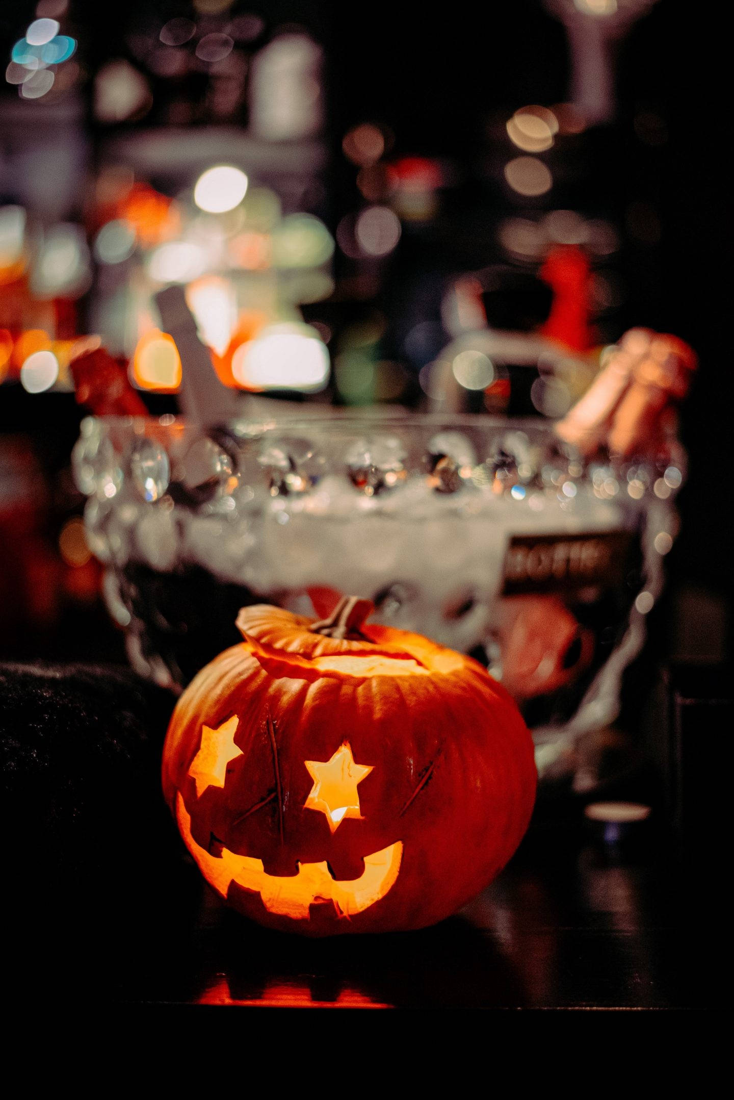 Spooky Aesthetic 桌布 壁紙 — 一個發光的傑克南瓜燈，帶有星形眼睛，放在酒吧櫃檯上，周圍是模糊的節日燈光。