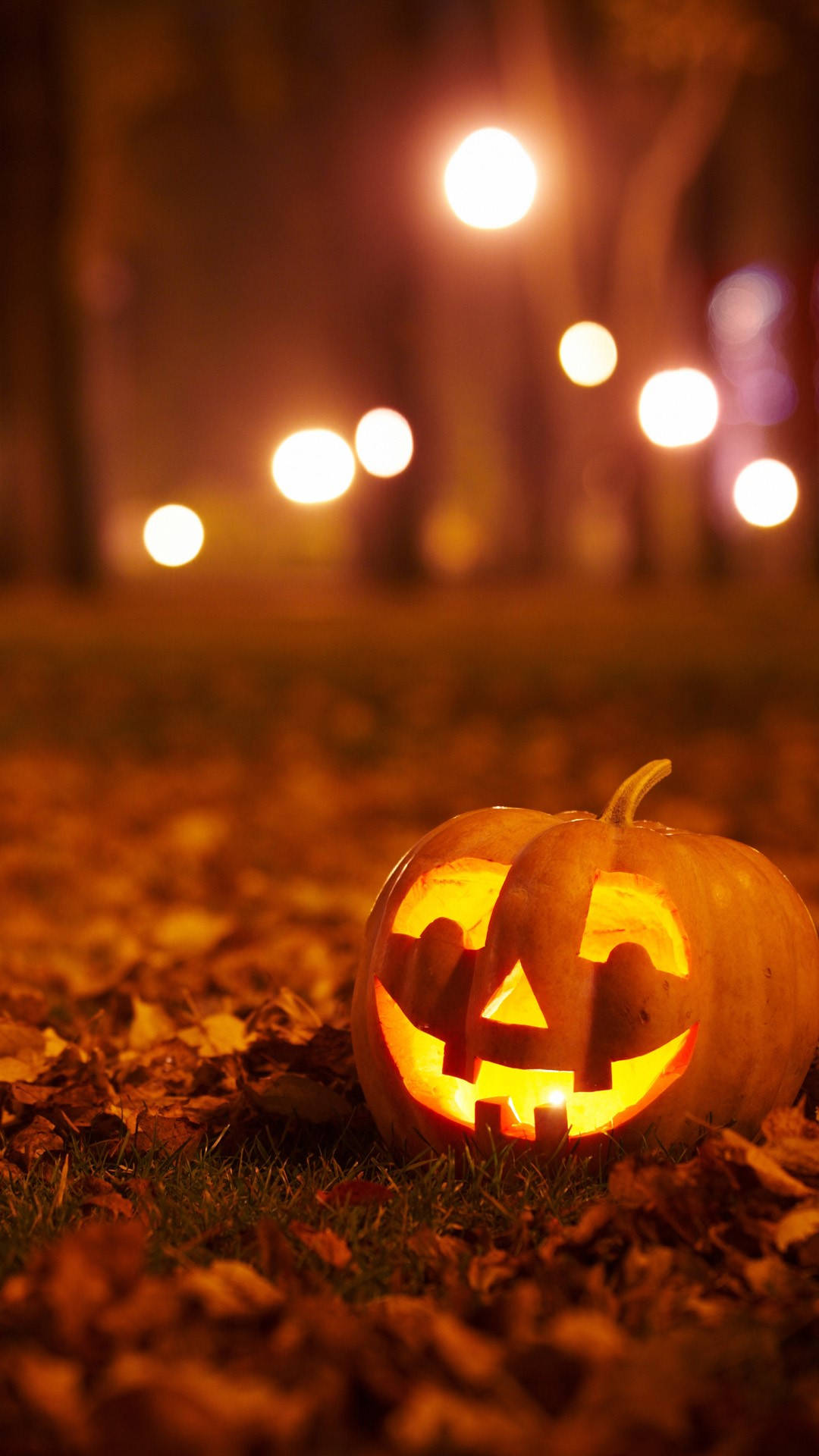 Halloween iPhone 桌布 壁紙 — 一個發光的傑克南瓜燈，帶有雕刻的笑容，在夜晚溫暖的散景街燈下坐在秋葉上。