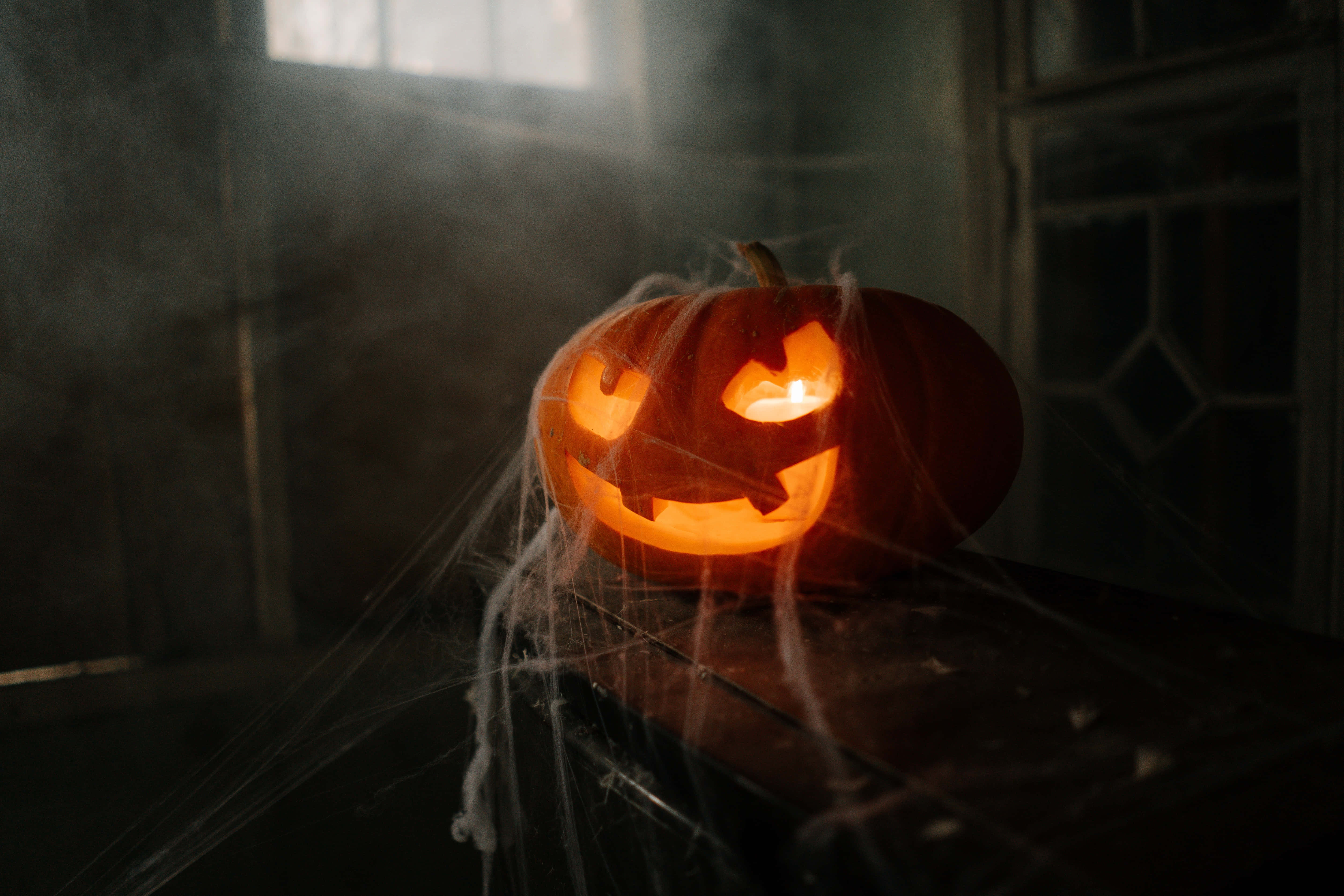 Pumpkin Patch 桌布 壁紙 — 一個發光的傑克南瓜燈，帶著陰險的笑容，放在佈滿灰塵、覆蓋蜘蛛網的表面上，在昏暗煙霧瀰漫的房間中投射出詭異的光芒。