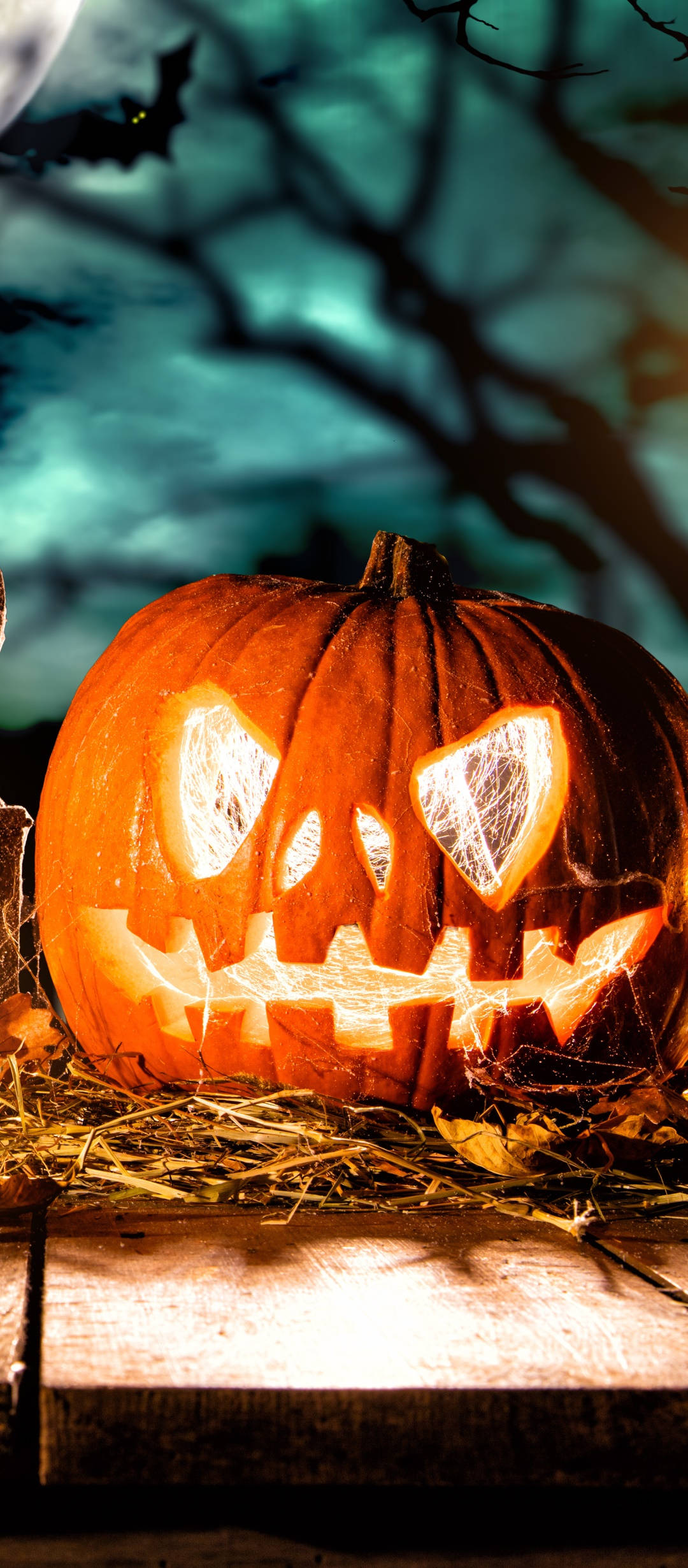 Cute Halloween Phone 桌布 壁紙 — 一個發光的傑克南瓜燈，有鋸齒狀的牙齒和心形眼睛，坐在乾草上，月光下的天空中有樹木剪影和一隻飛行的蝙蝠。