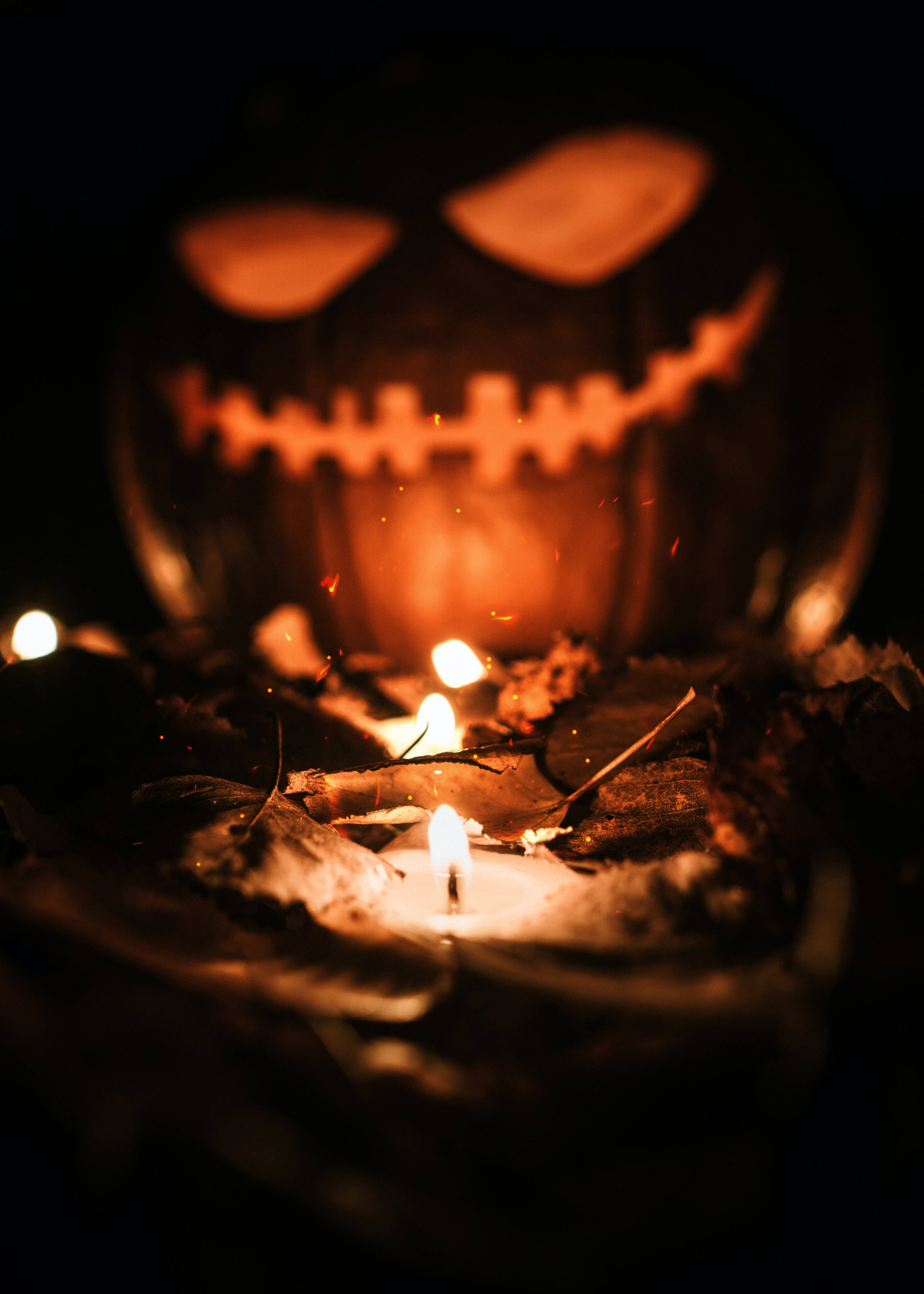Halloween Phone 桌布 壁紙 — 一個發光的傑克南瓜燈，縫合的嘴巴，在黑暗中的閃爍蠟燭和落葉後面若隱若現。