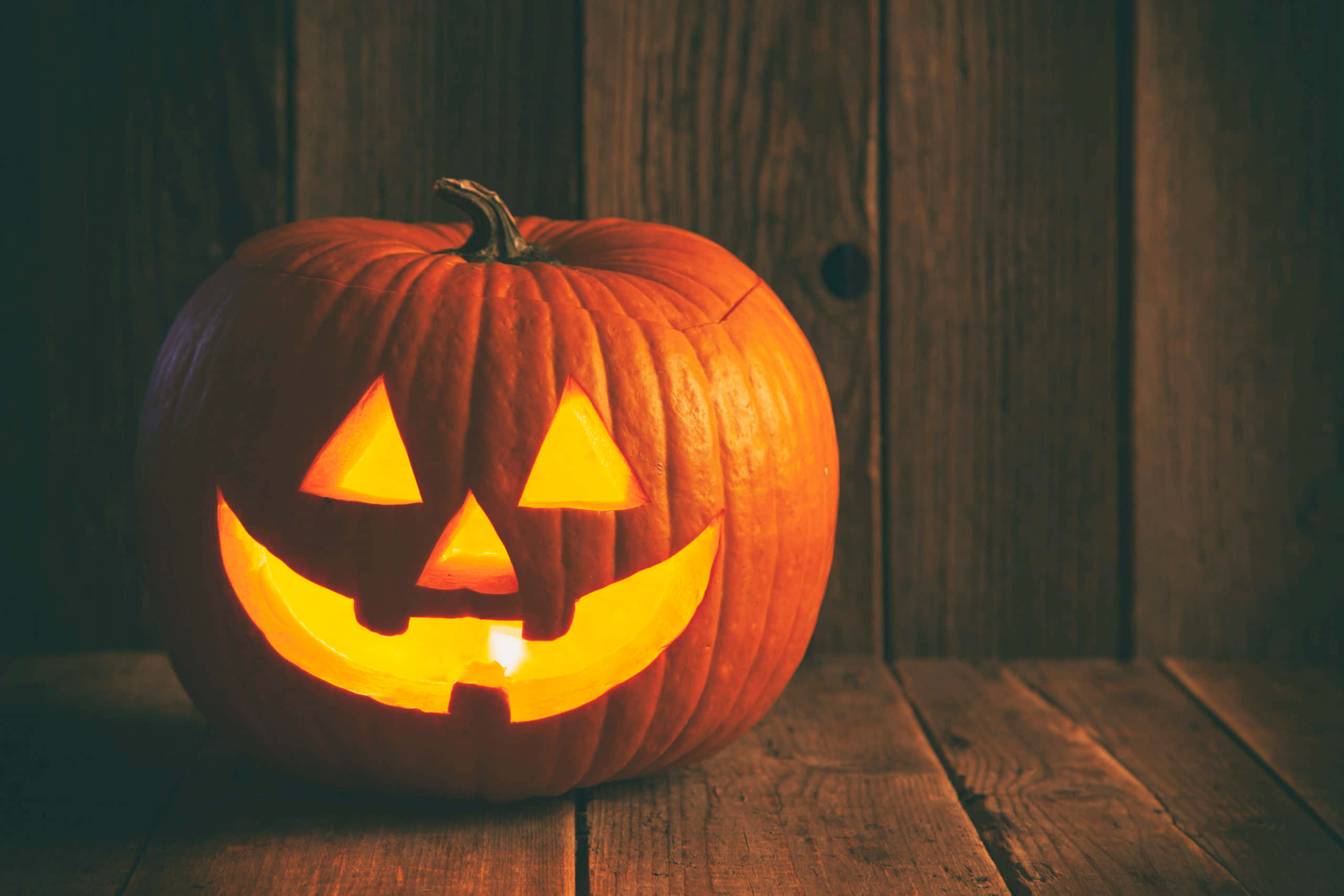 Jack O Lantern 桌布 壁紙 — 一個發光的傑克南瓜燈，雕刻著咧嘴笑，放在質樸的木桌上，背景是深色木牆。