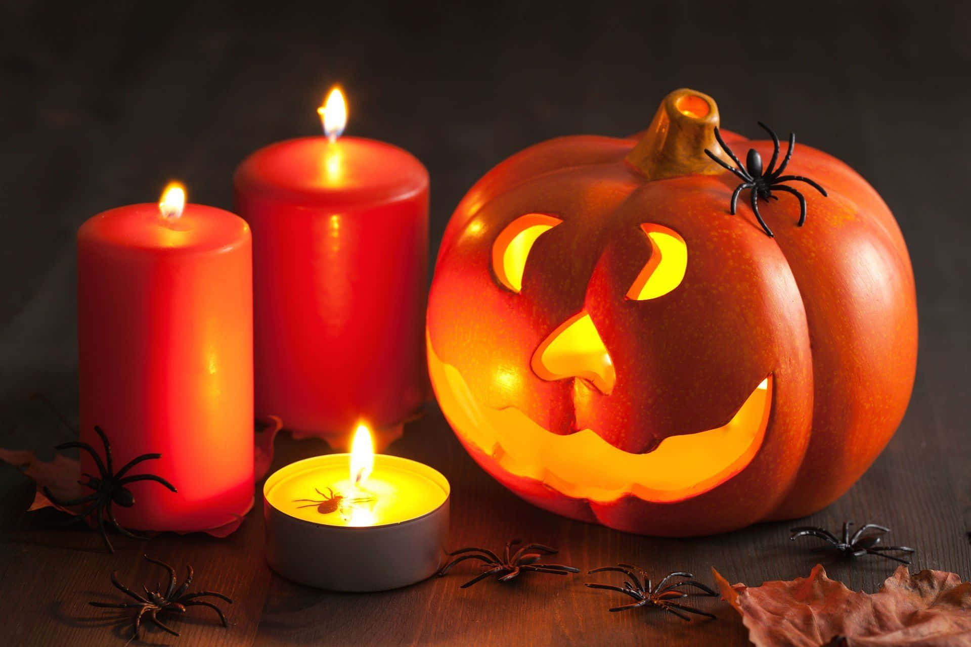 Halloween Candles 桌布 壁紙 — 一個發光的傑克南瓜燈，雕刻著臉孔，旁邊有三根點燃的紅色蠟燭和散落的塑膠蜘蛛，放在深色木桌上。