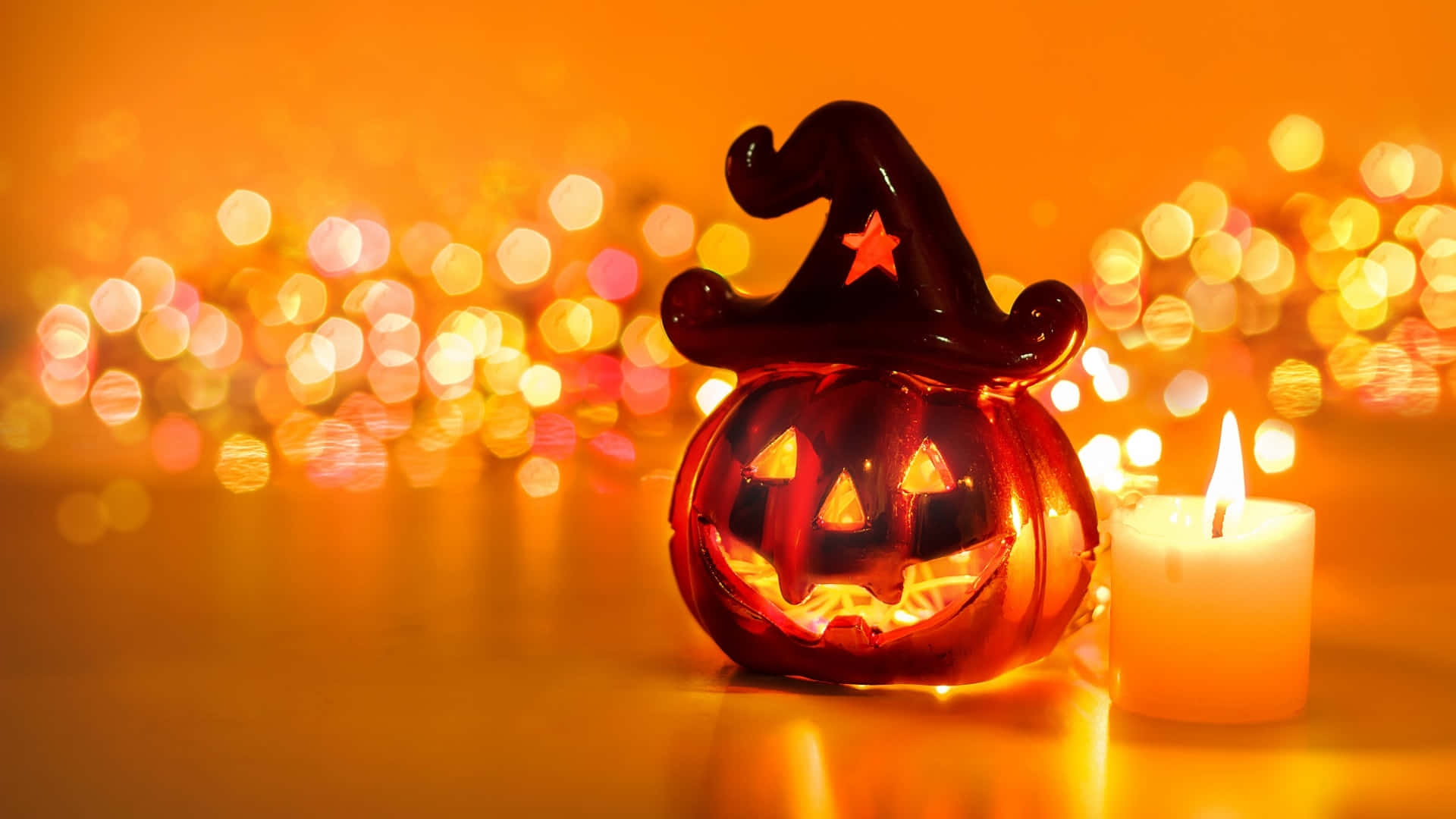 Orange Halloween 桌布 壁紙 — 一個發光的傑克南瓜燈戴著女巫帽，坐在點燃的蠟燭旁邊，背景是溫暖的散景。