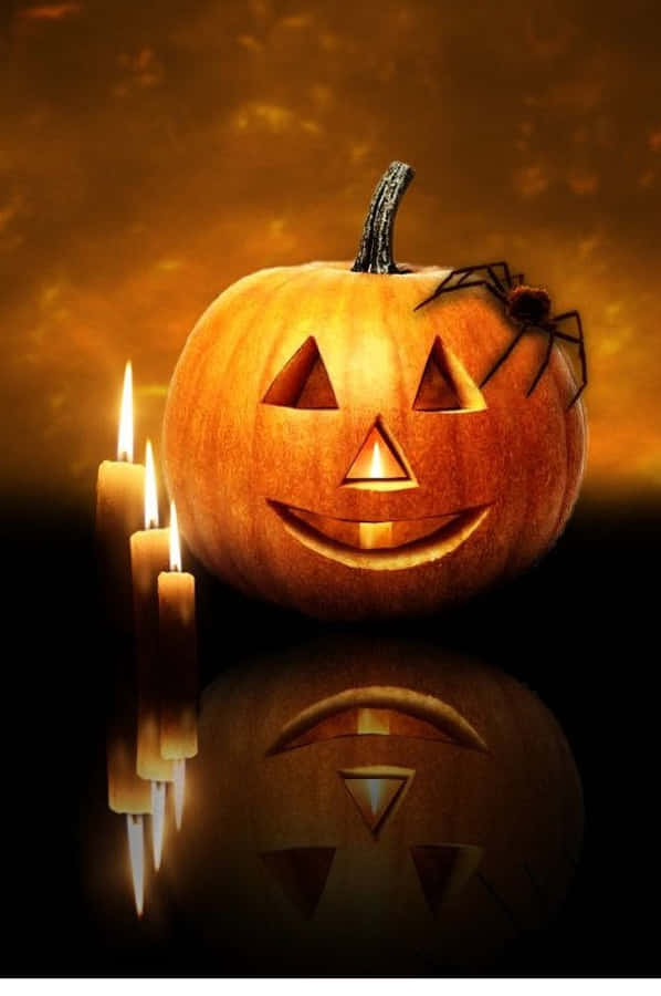 iPhone Halloween 桌布 壁紙 — 一個發光的南瓜燈，側面有一隻蜘蛛，旁邊有三根點燃的蠟燭，背景是溫暖的橙色。