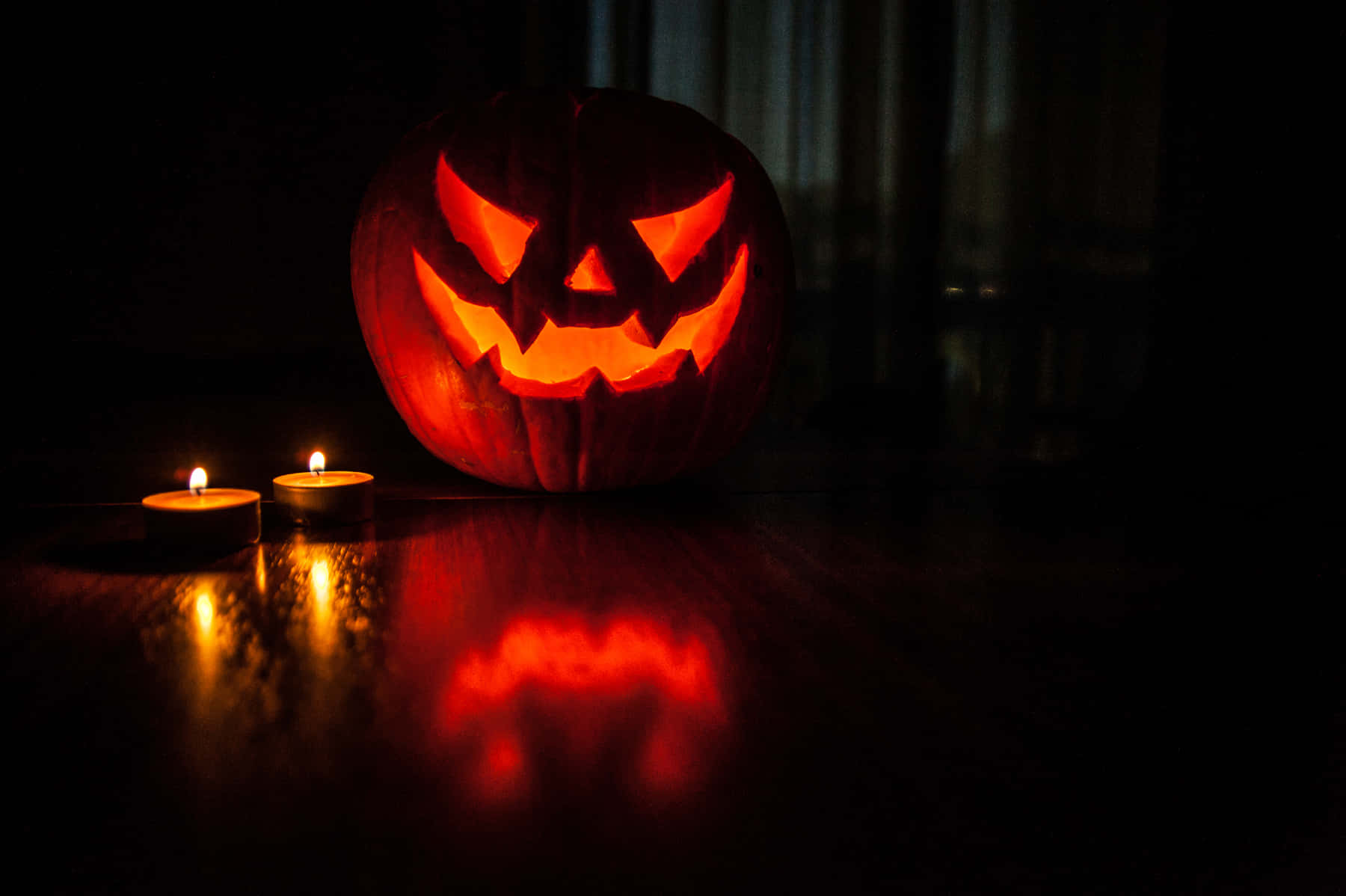 Creepy Halloween 桌布 壁紙 — 一個發光的南瓜燈，帶著猙獰的笑容，放在深色木質表面上，旁邊有兩根點燃的蠟燭，投射出詭異的倒影。