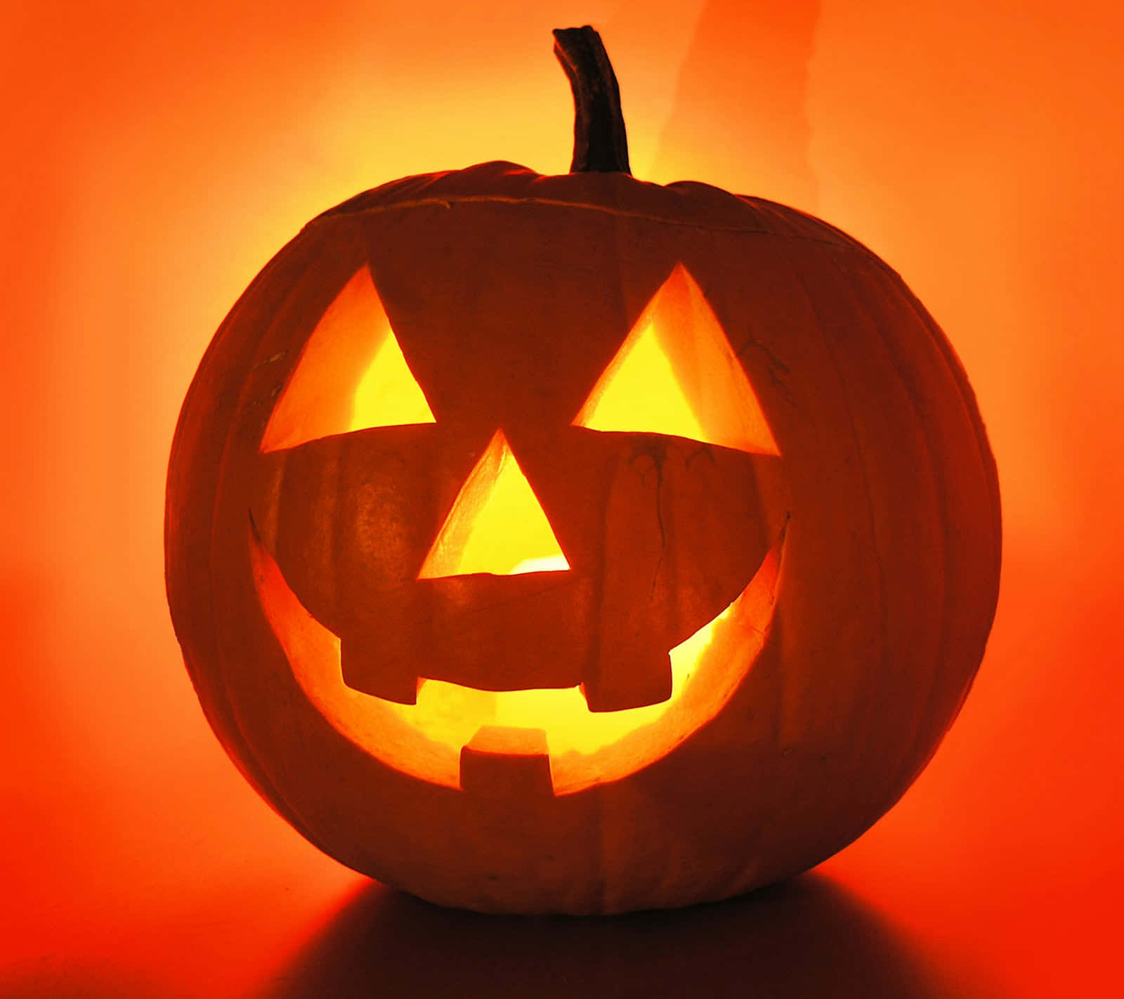 Jack O Lantern 桌布 壁紙 — 一個發光的南瓜燈，有著三角形眼睛和露齒笑容，在純橙色背景上散發出溫暖的橙色光芒。
