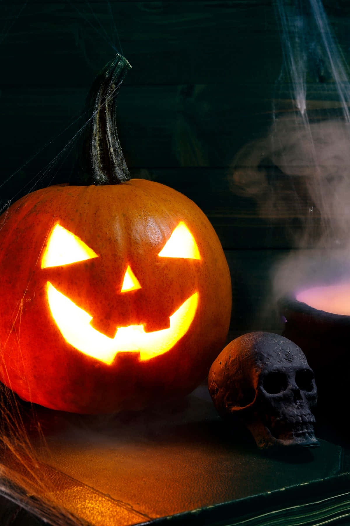 Creepy Halloween 桌布 壁紙 — 一個發光的南瓜燈帶著邪惡的笑容，坐在一個骷髏頭旁邊，位於深色木質表面上，周圍環繞著薄霧和蜘蛛網。