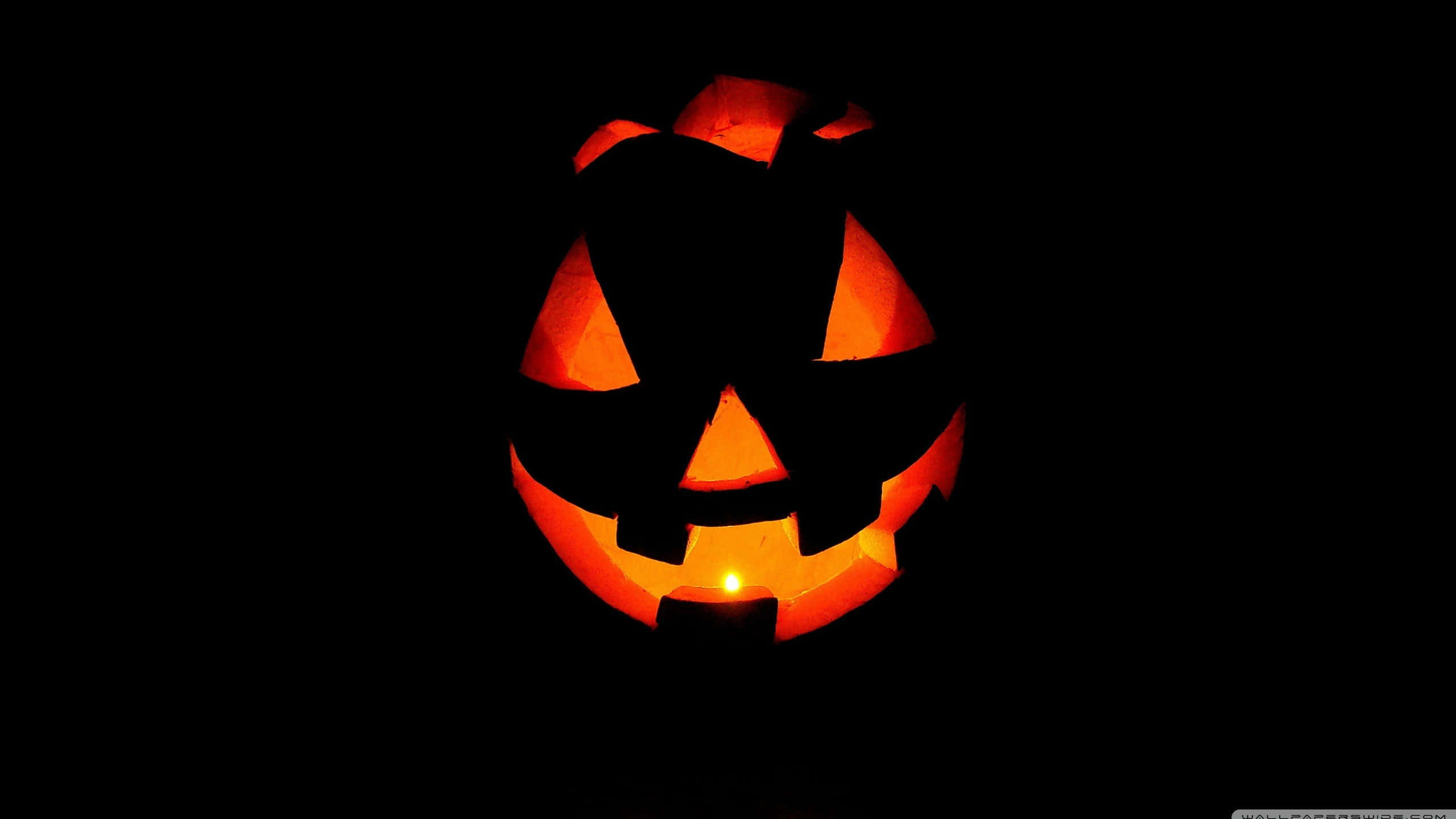 3d Halloween 桌布 壁紙 — 一個發光的南瓜燈籠，帶有三角形眼睛和鋸齒狀笑容，由蠟燭照亮，背景為黑色。