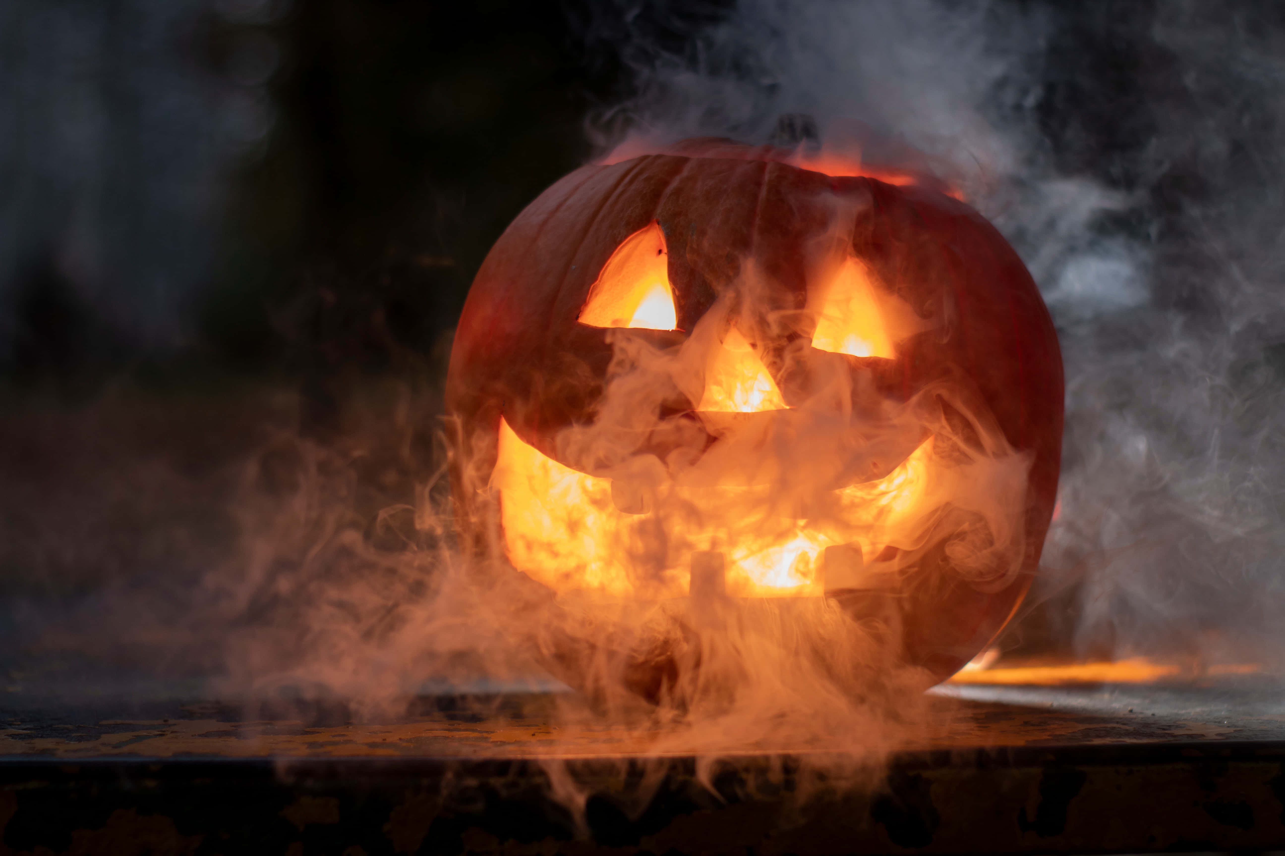 Halloween MacBook 桌布 壁紙 — 一個發光的南瓜燈籠，帶著兇險的笑容，在黑暗背景下散發出火焰般的光芒和旋轉的煙霧。