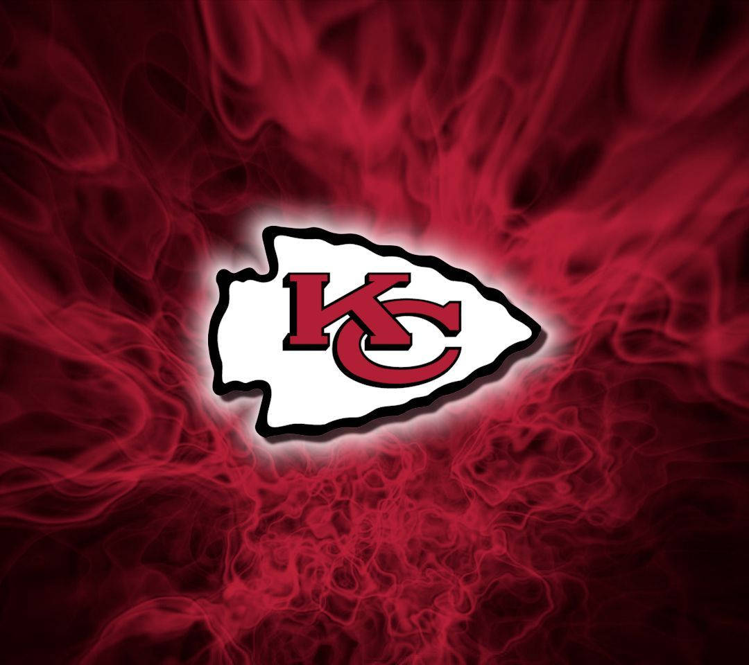 Kc Chiefs 桌布 壁紙 — 一個發光的堪薩斯城酋長隊標誌從旋轉的紅色煙霧中浮現，背景為深色。