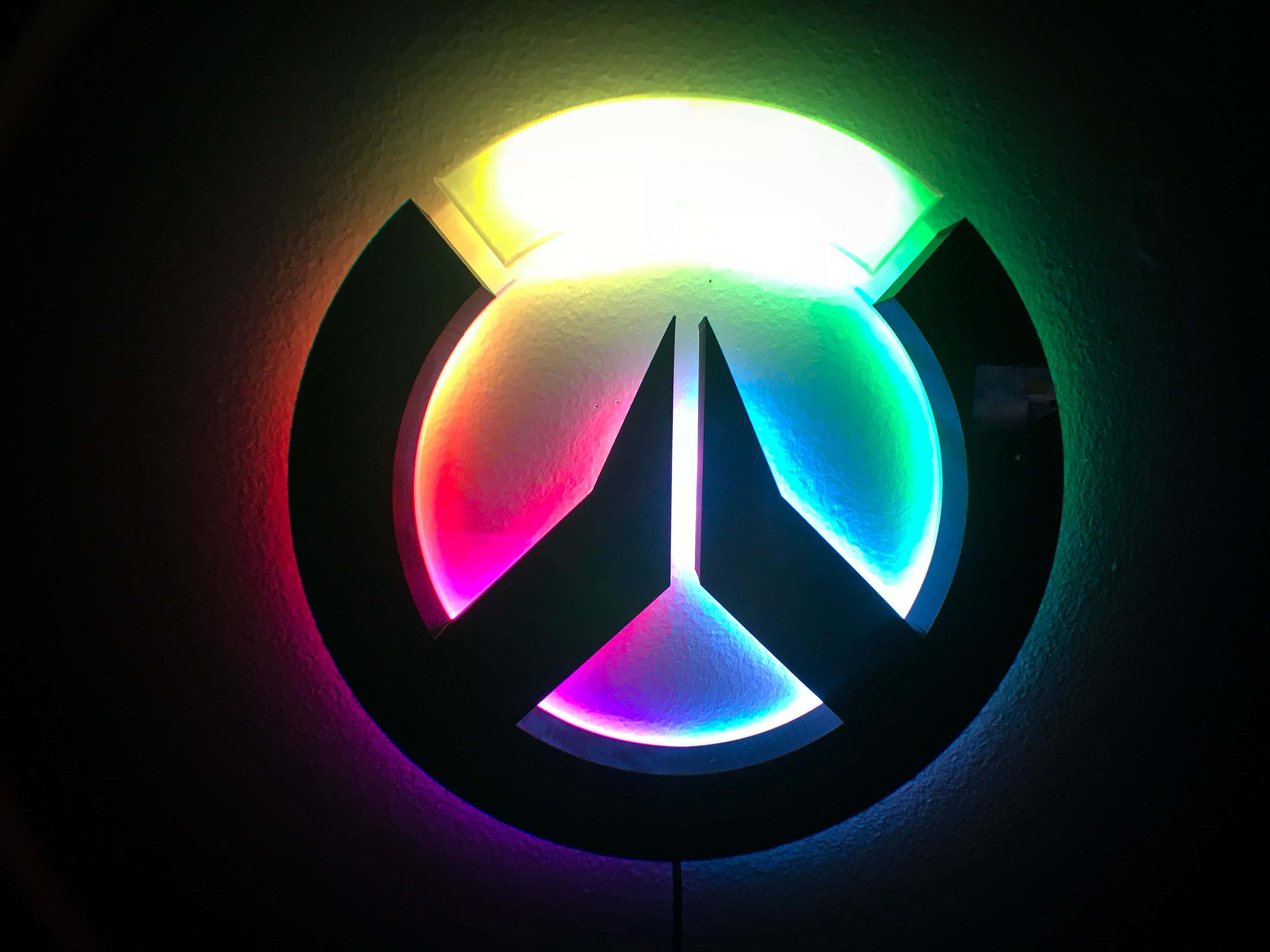 Aesthetic Overwatch 桌布 壁紙 — 一個發光的彩虹色《鬥陣特攻》標誌安裝在深色牆壁上，在昏暗的房間中投射出鮮豔的彩色光線。
