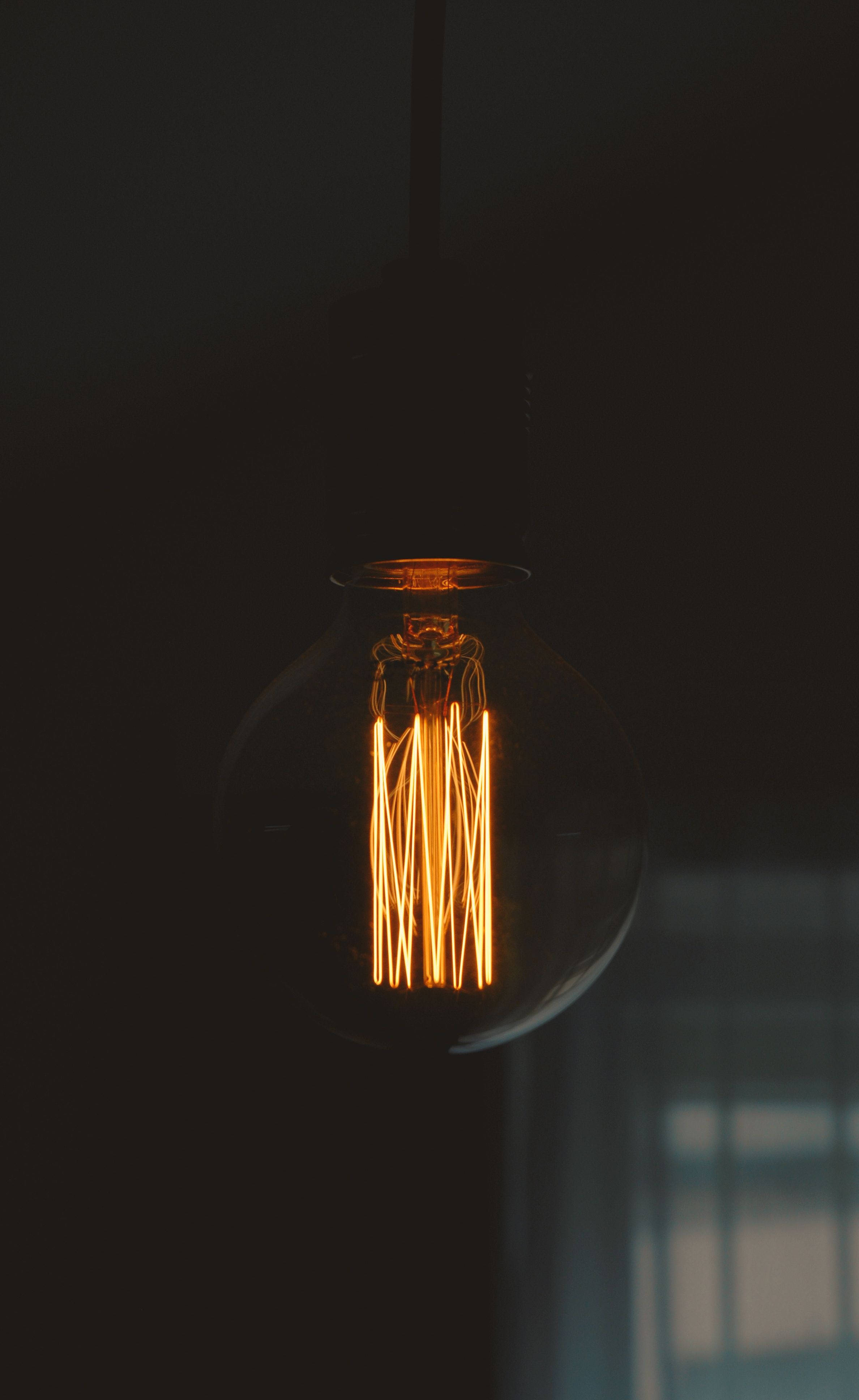 Light Bulb 桌布 壁紙 — 一個發光的復古燈絲燈泡懸掛在黑暗中，透過玻璃外殼散發出溫暖的金色光芒。