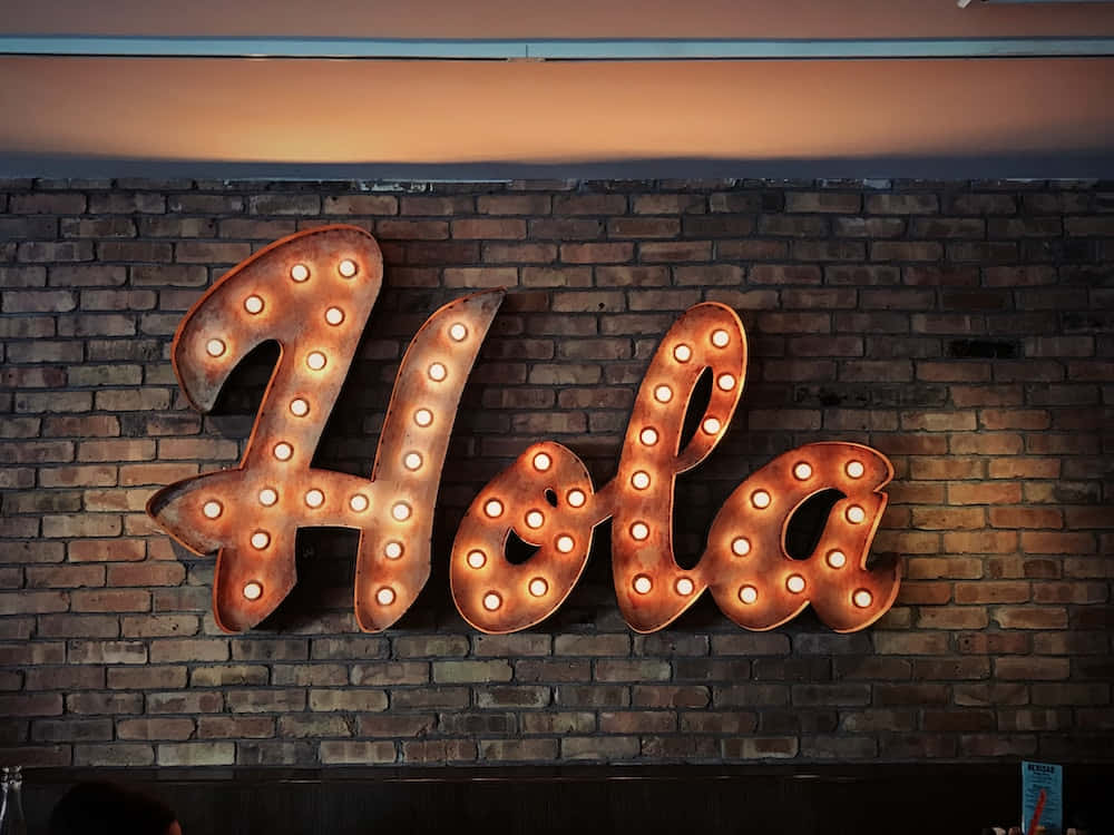 Spanish 桌布 壁紙 — 一個發光的招牌，用草書字母拼出「Hola」，帶有溫暖的燈泡，安裝在質樸的磚牆上。