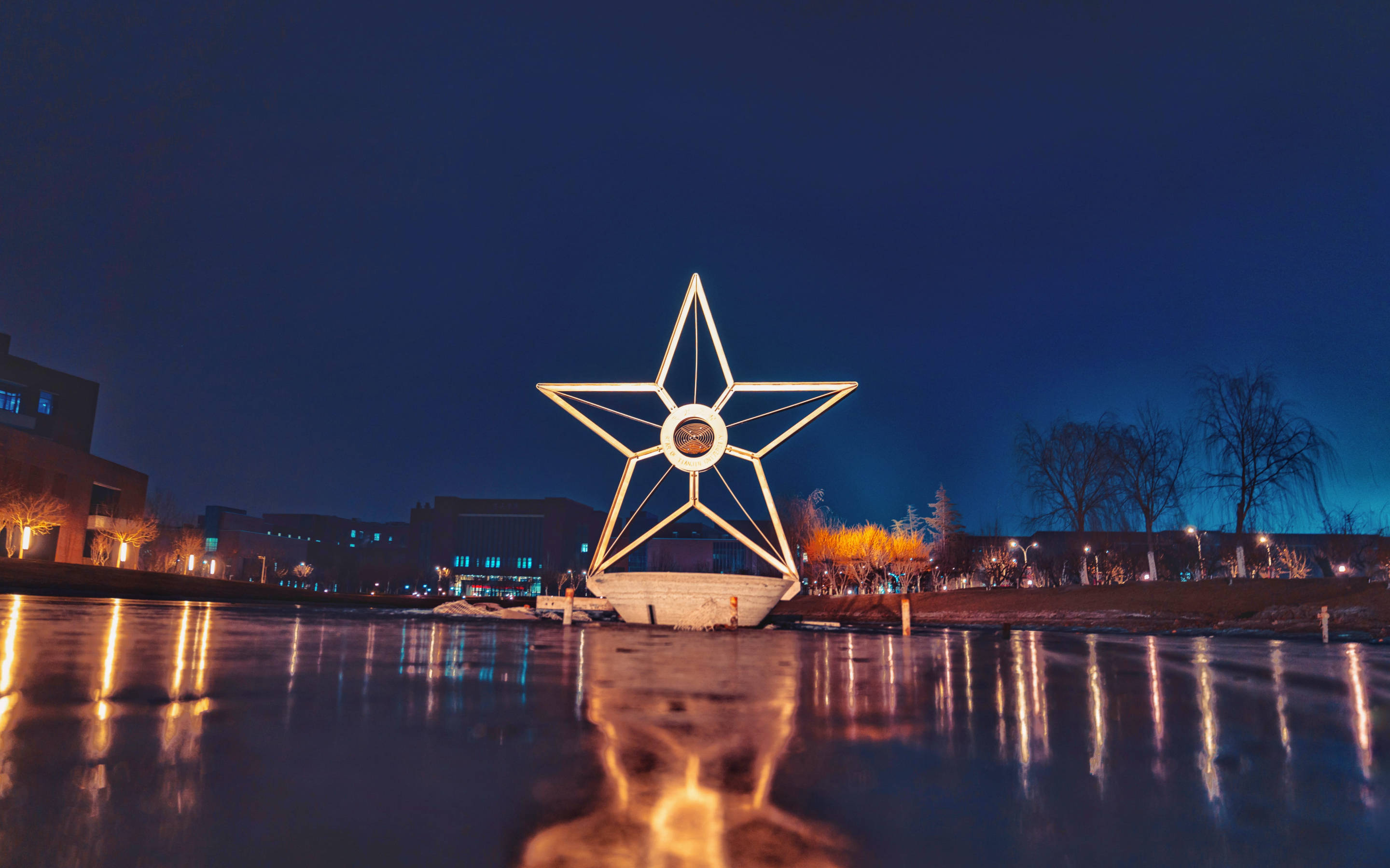 Tianjin 桌布 壁紙 — 一個發光的星星雕塑在冰冷的湖水中倒映，背景是深藍色的夜空、光禿的樹木和遠處的建築物。