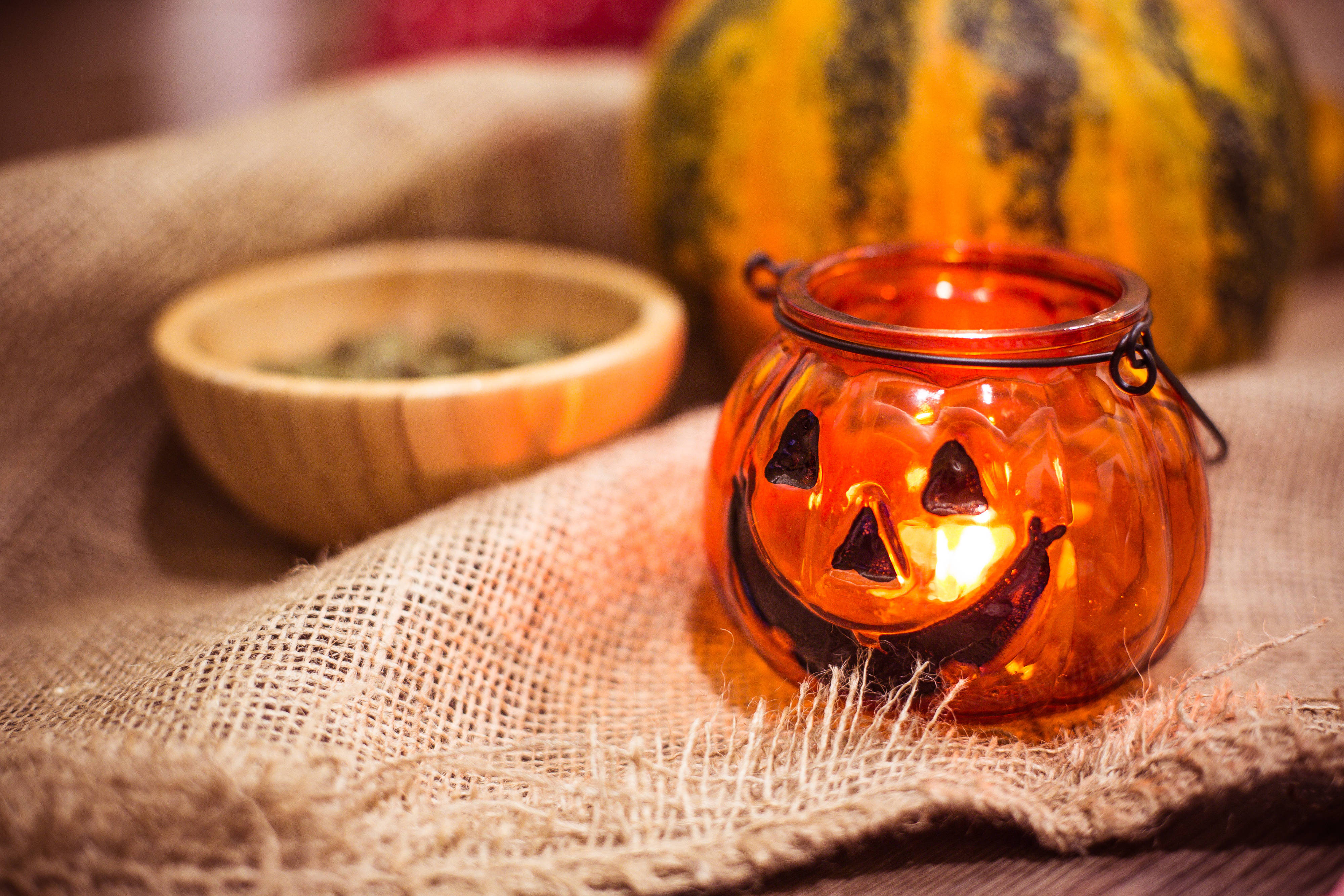 Cute Halloween Desktop 桌布 壁紙 — 一個發光的橙色南瓜燈燭罐放在粗麻布上，旁邊有一個木碗和條紋南瓜。