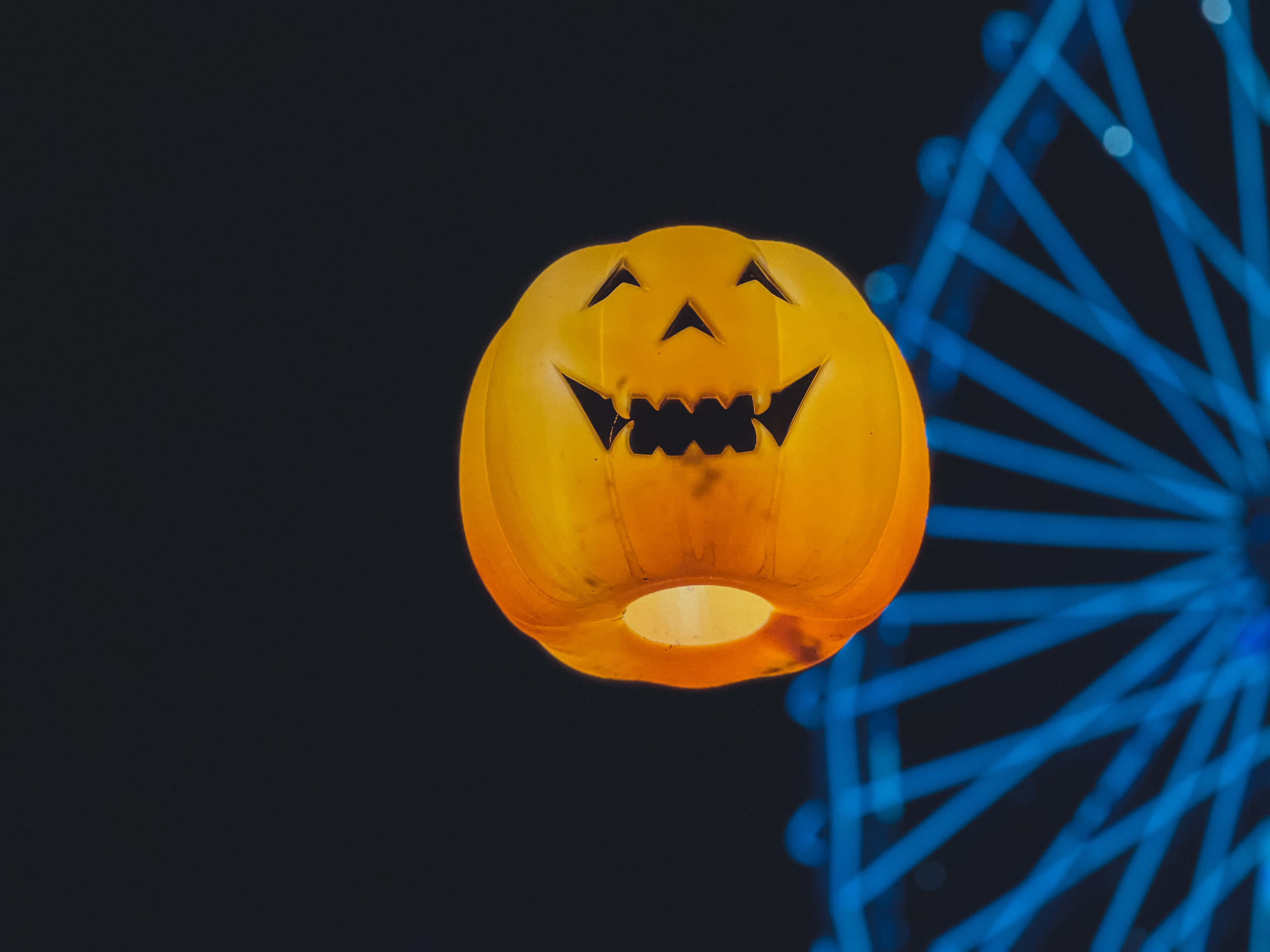 jack o'lantern 桌布 壁紙 — 一個發光的橙色南瓜燈籠，帶有雕刻的笑容，在黑暗的夜空中漂浮在模糊的藍色摩天輪旁邊。