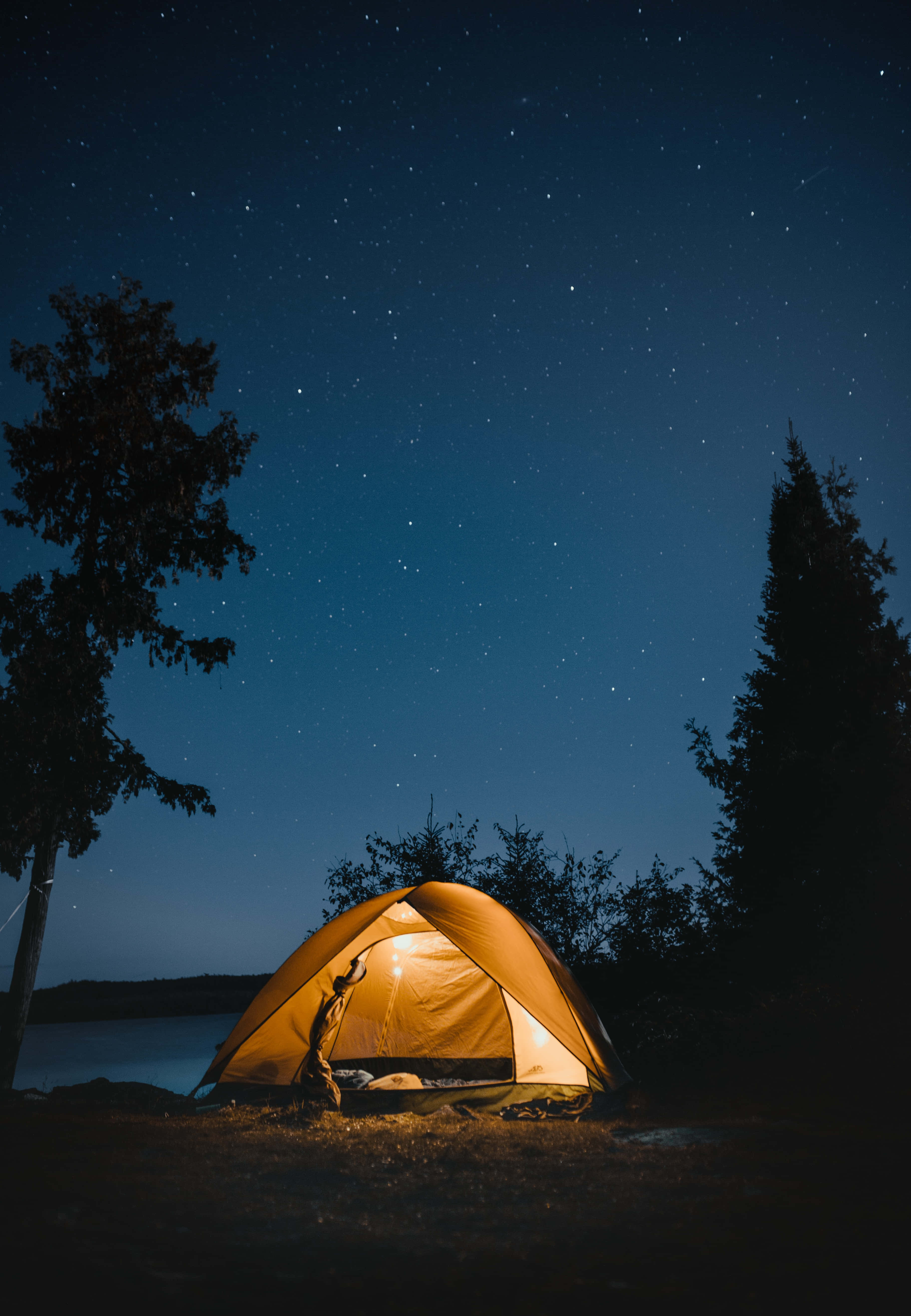Camping 桌布 壁紙 — 一個發光的橙色帳篷，坐落在湖邊，星空下，被黑暗的樹木環繞。