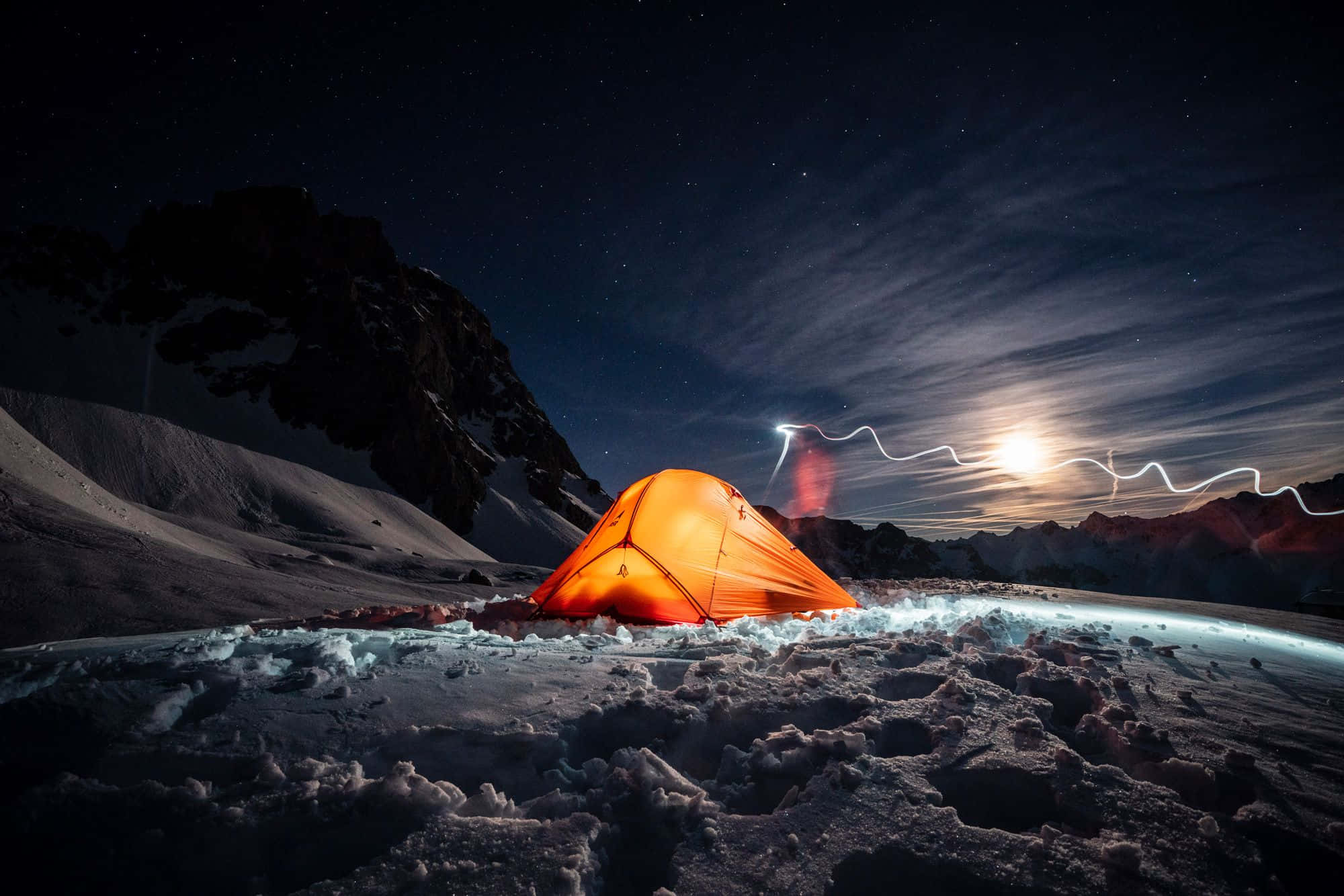 Winter Camping 桌布 壁紙 — 一個發光的橙色帳篷坐落在白雪皚皚的山脈中，在繁星點點的夜空下，戲劇性的閃電照亮了地平線。