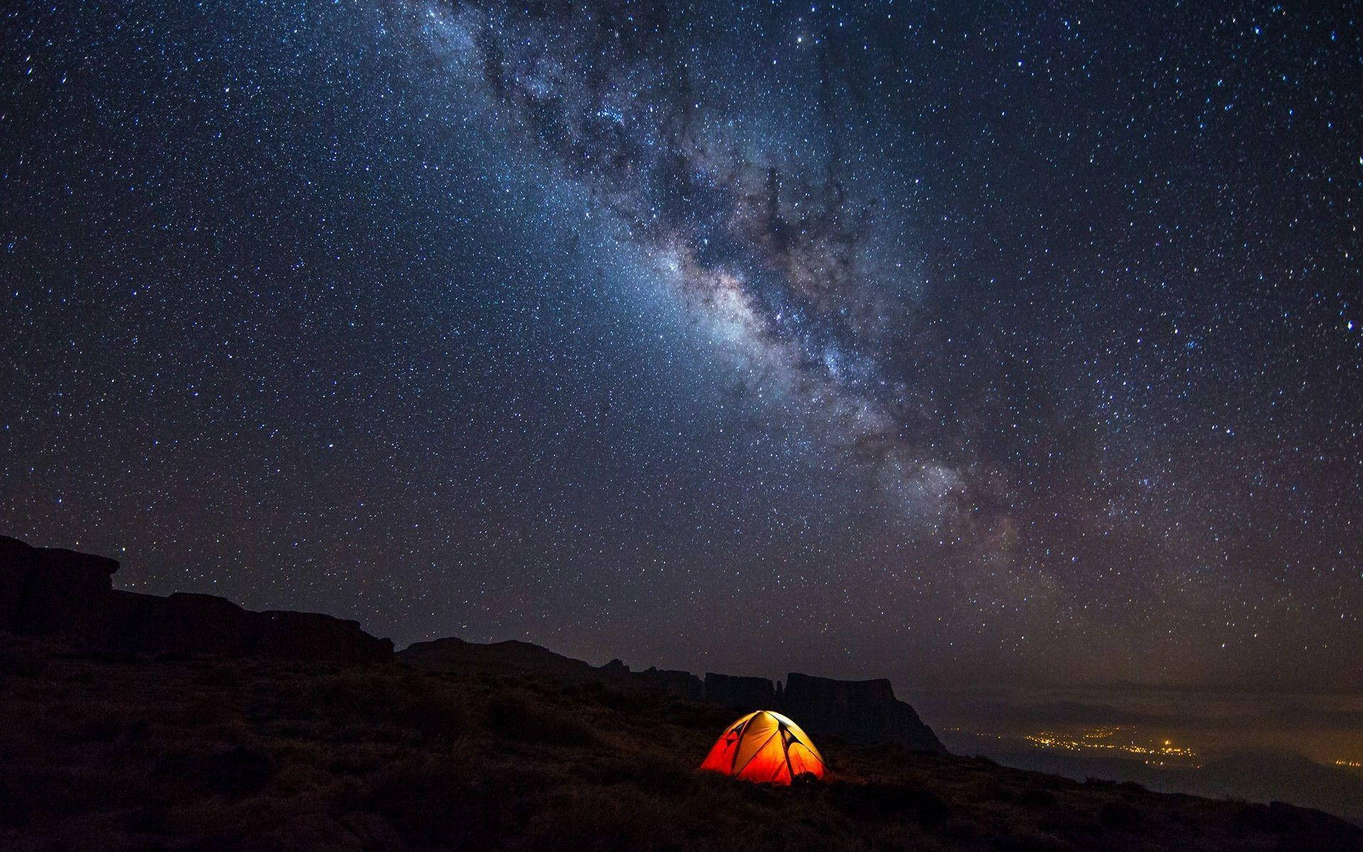 Camping 桌布 壁紙 — 一個發光的橙色帳篷獨自矗立在岩石山坡上，下方是令人嘆為觀止的星空，充滿了銀河。
